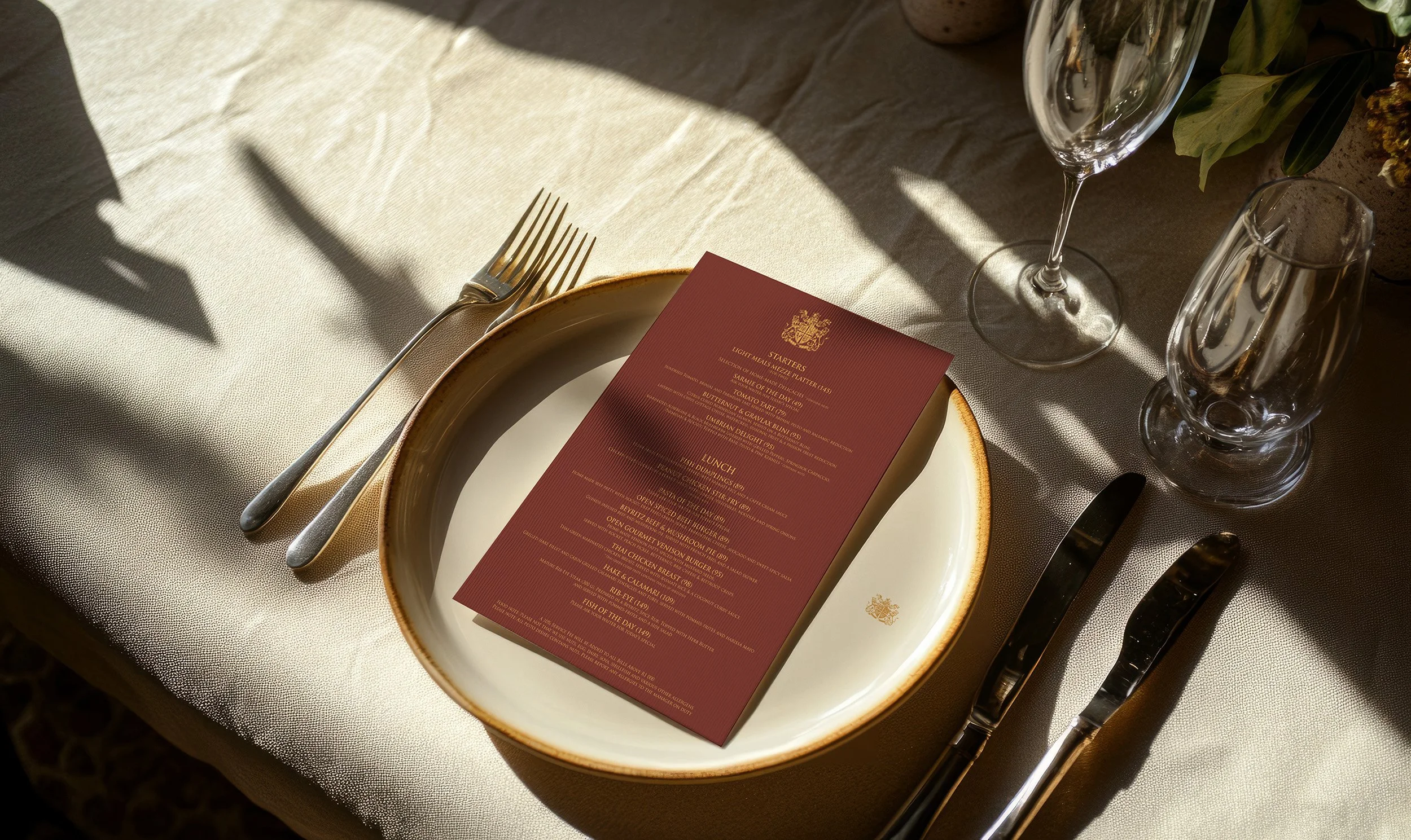 Table setting with a beige tablecloth, a white plate with a gold rim, a red menu on top, and silverware including a fork, knife, and spoon. There are also a champagne flute and a water glass, with part of a floral arrangement visible.