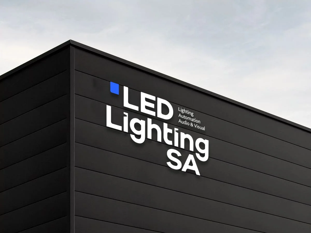 Close-up of a modern building with black exterior siding, featuring a sign that reads 'LED Lighting SA' with smaller text underneath that says 'Lighting Automation Audio & Visual', and a small blue square symbol at the top left of the sign.