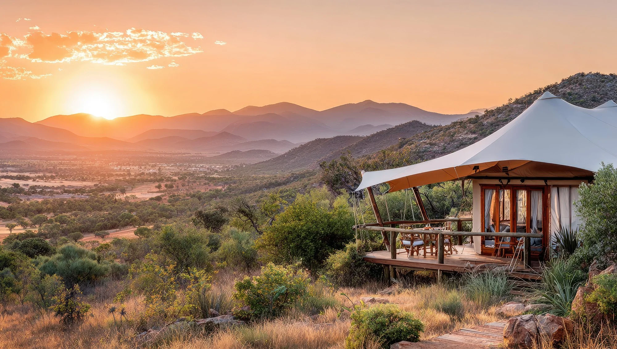 A tented accommodation on a hillside with a sunrise over a mountainous landscape, surrounded by desert vegetation.