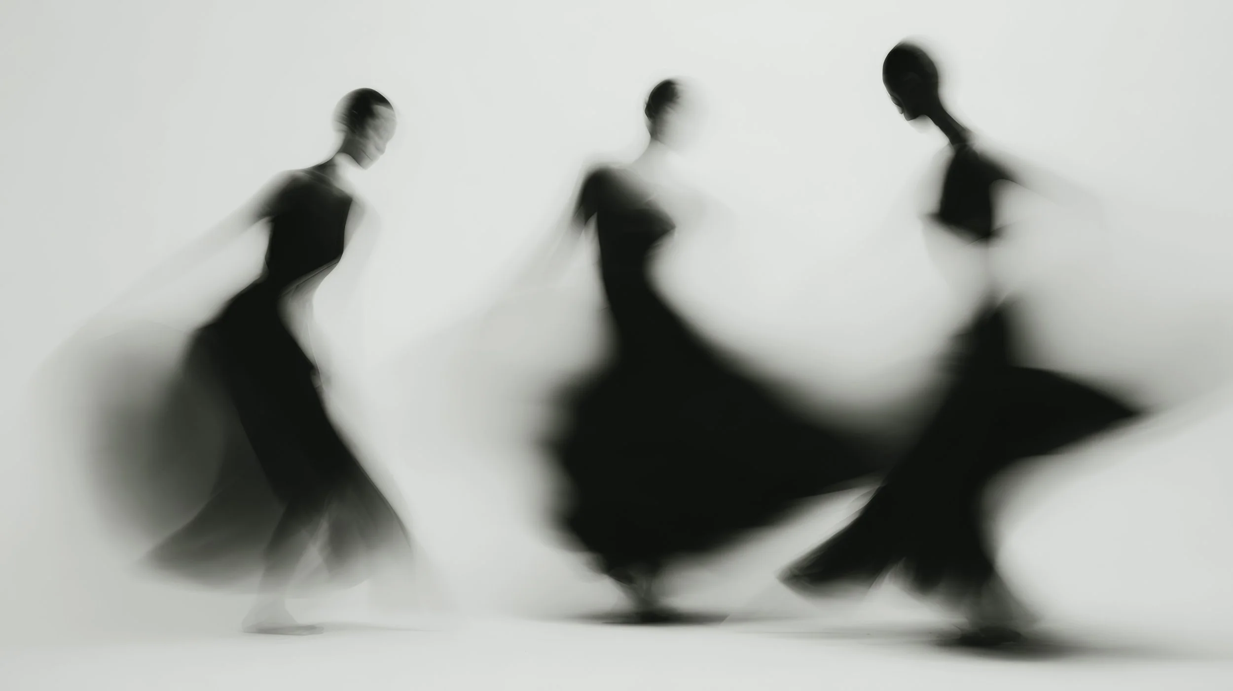 Three blurred figures of women in black dresses holding hands, viewed from below and backlit, creating a high-contrast, abstract silhouette.
