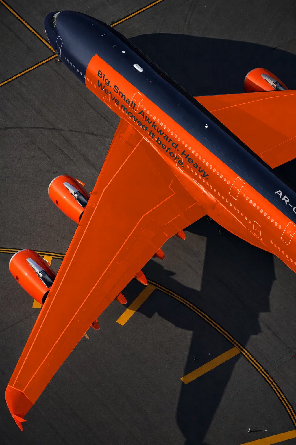An airplane on the tarmac with a black and orange color scheme. The fuselage has a message that reads, "Big. Small. Awkward. Heavy. We’ve moved it before." The airplane's shadow is visible on the ground.