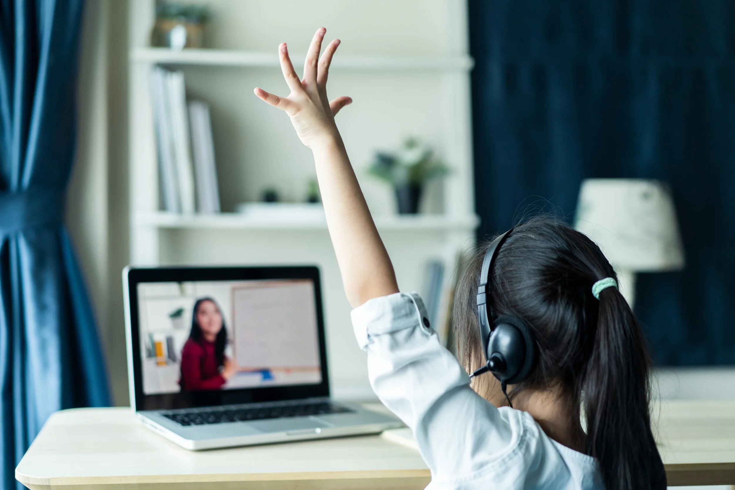 A young girl in headphones raising her hand during an online class with a teacher on her laptop.