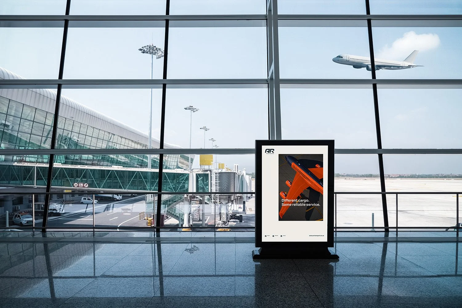 View of an airport terminal with large glass windows, showing airplanes and airport structures outside. Inside, a digital advertisement display features an orange cargo plane with the text "Different cargo. Same reliable service.".