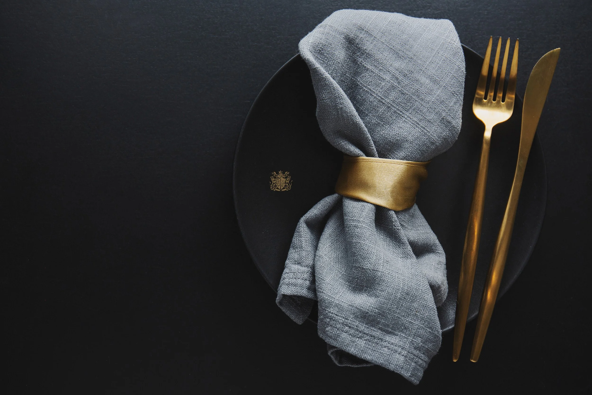 Elegant black plate with a gold emblem, grey cloth napkin tied with a gold ribbon, and gold fork and knife on a black surface.