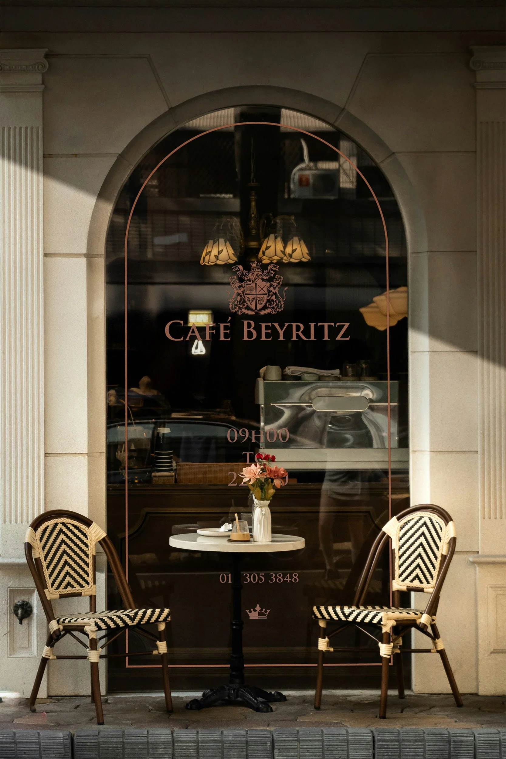 The storefront of Café Beyritz with a large glass window showing inside lighting fixtures, a table with a vase of flowers, and two wicker chairs outside. The café's signs display the name, opening hours, and contact information.