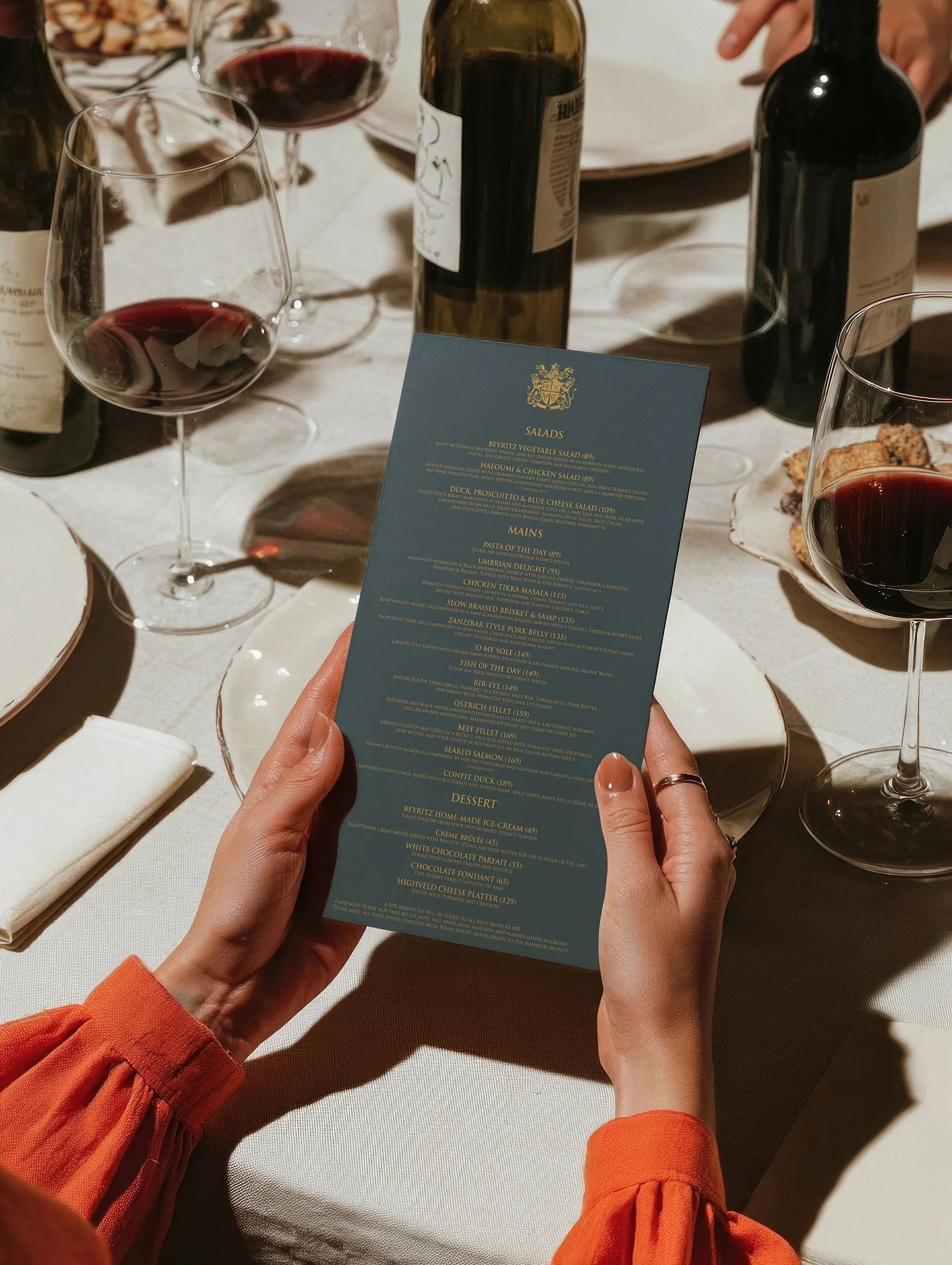 Person holding a blue dinner menu at a table with glasses of red wine and bottles, with a white tablecloth.