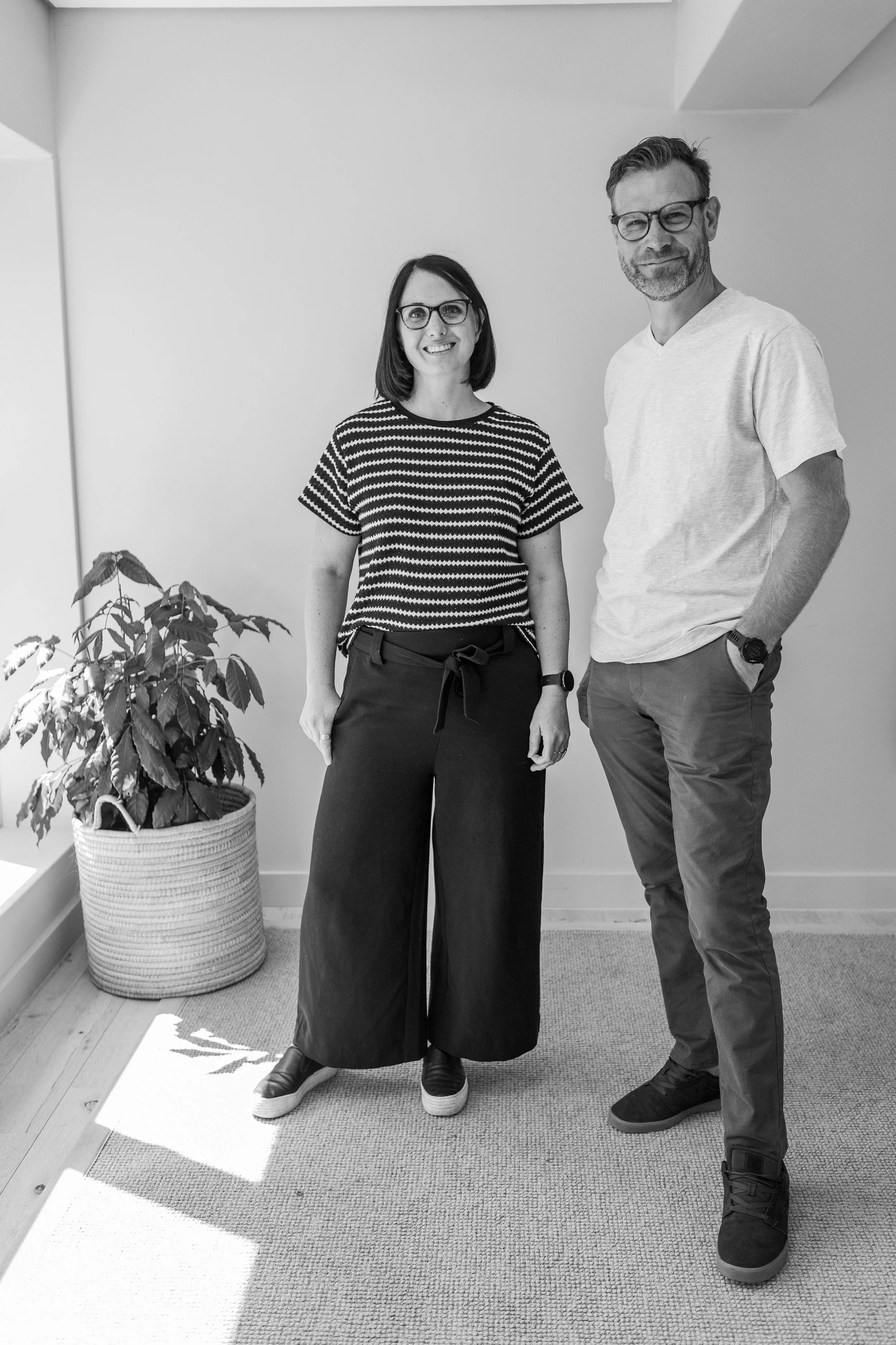 A woman and a man standing indoors next to a potted plant, both smiling and wearing glasses.