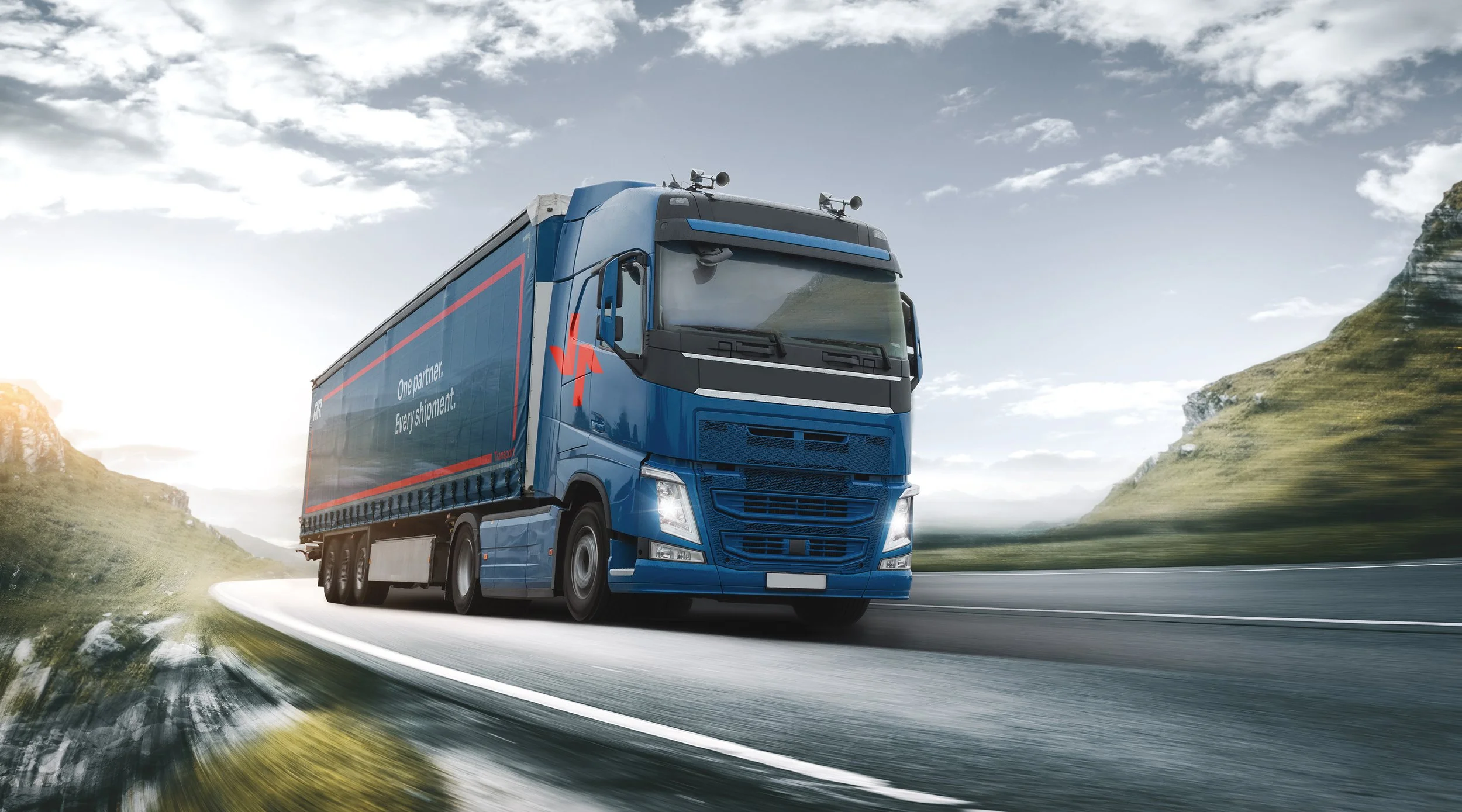A blue semi-truck driving on a winding mountain road during daytime, with green hills and a partly cloudy sky in the background.