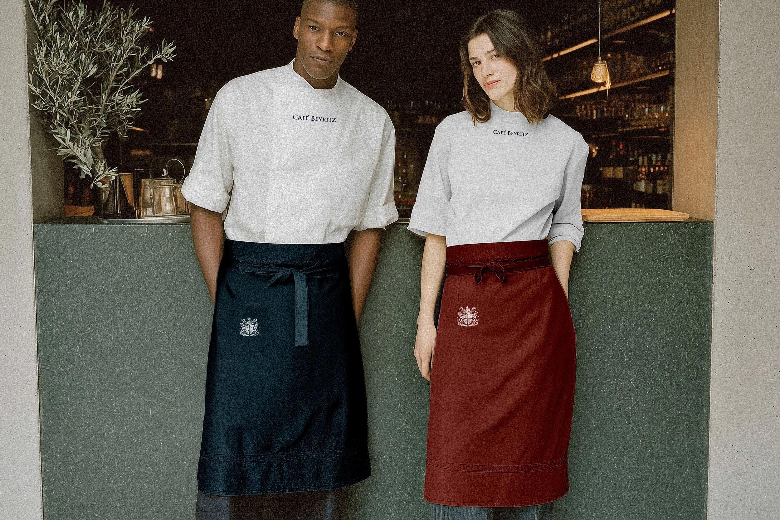 Two restaurant staff members standing behind a bar counter; man wearing a white chef's coat with 'Cafe Beyritz' embroidered on it and a dark apron, woman wearing a white shirt with 'Cafe Beyritz' and a red apron, both with arms at their sides, in a dimly lit restaurant or bar setting.