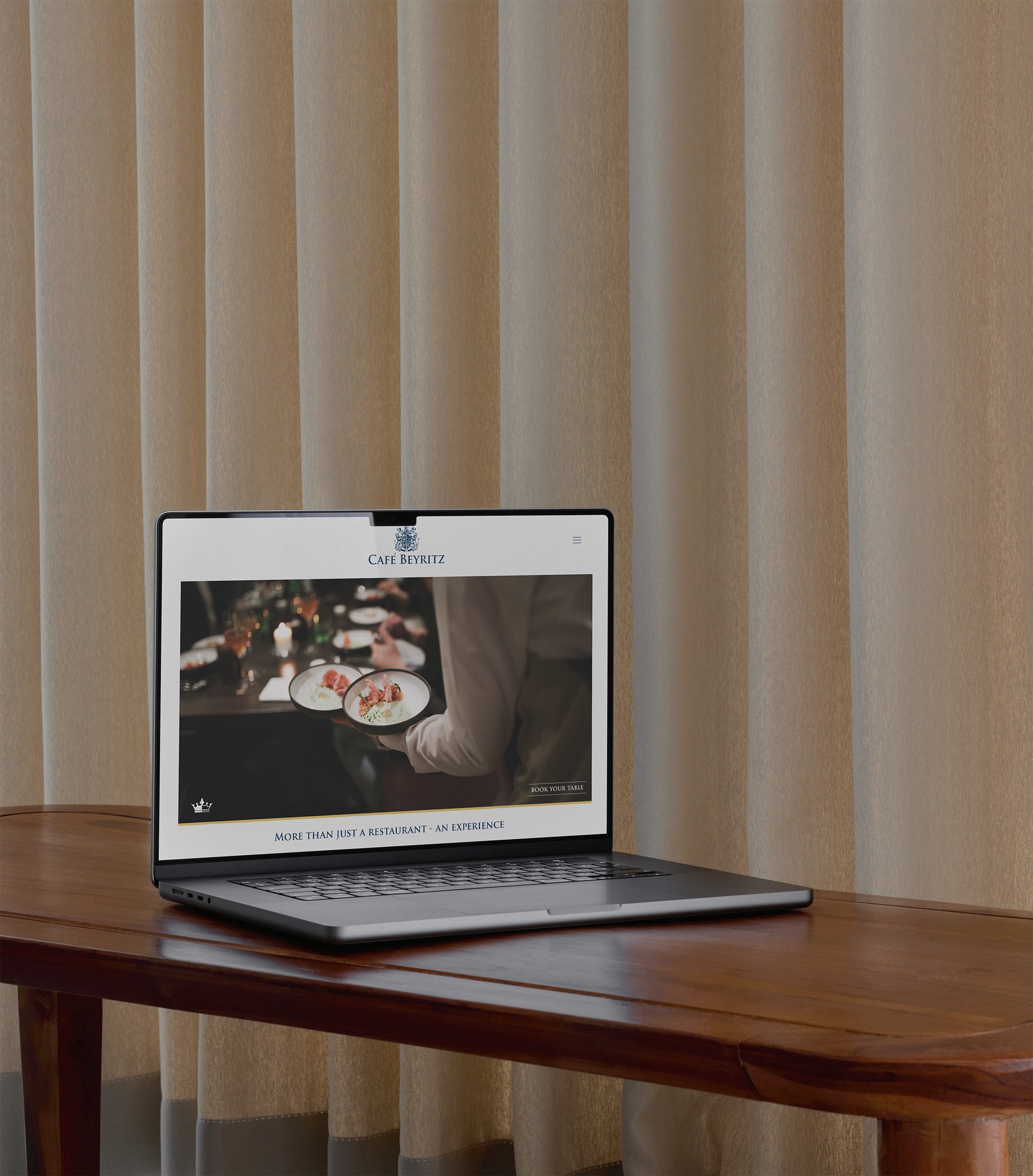 A laptop on a wooden table displaying a website for Cafe Beyritz with an image of a waiter serving food, and a blurred background of a dining setting with candles and plates.