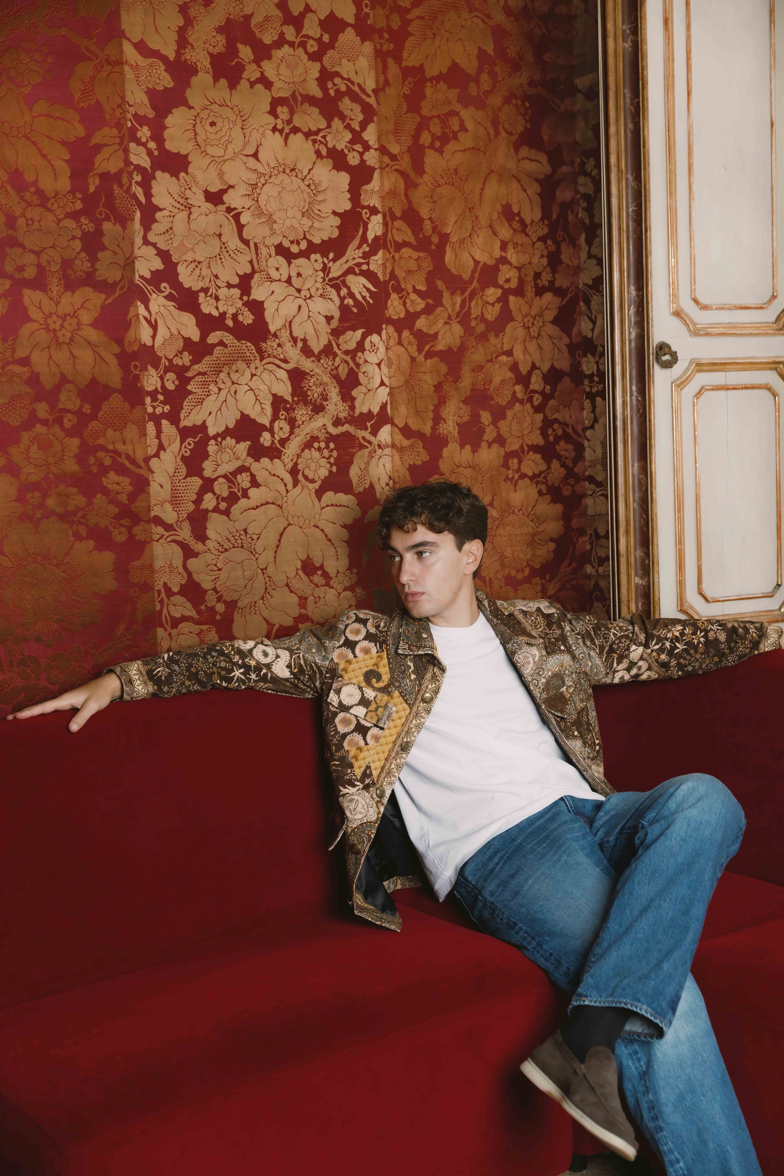 Giovane uomo seduto su un divano rosso con sfondo di carta da parati rossa e dorata, che indossa una camicia decorata, una maglietta bianca e jeans.