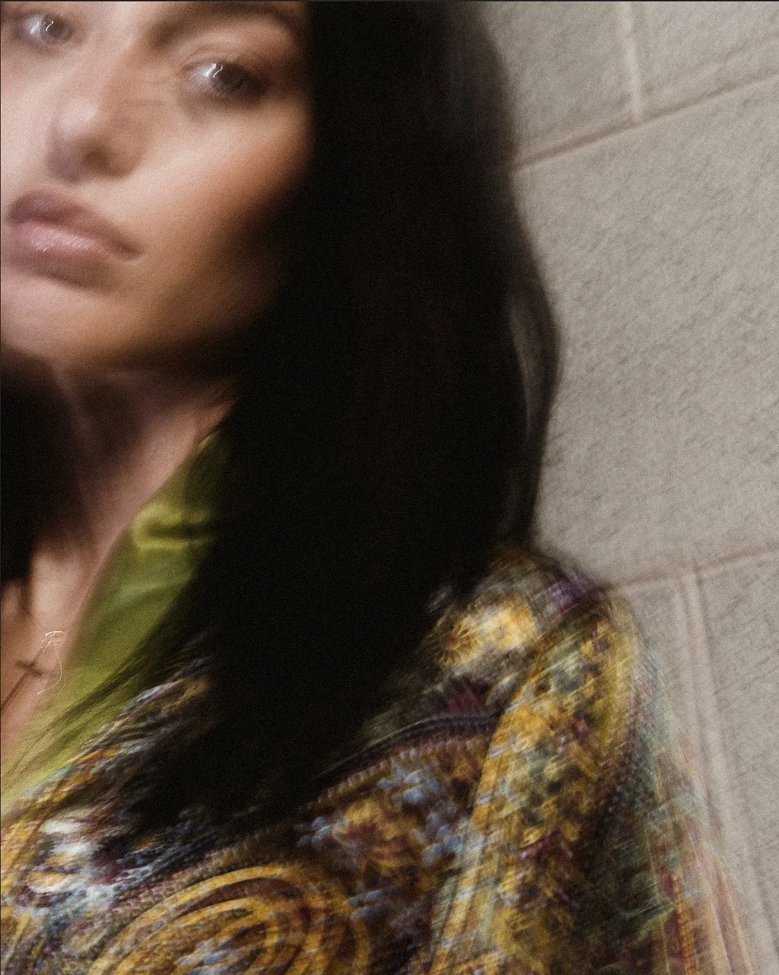 Ragazza con capelli neri, occhi grandi, labbra sottili e pelle chiara, che indossa un abito colorato con motivi intricati. Sembra essere in un ambiente interno con pareti di mattoni beige.