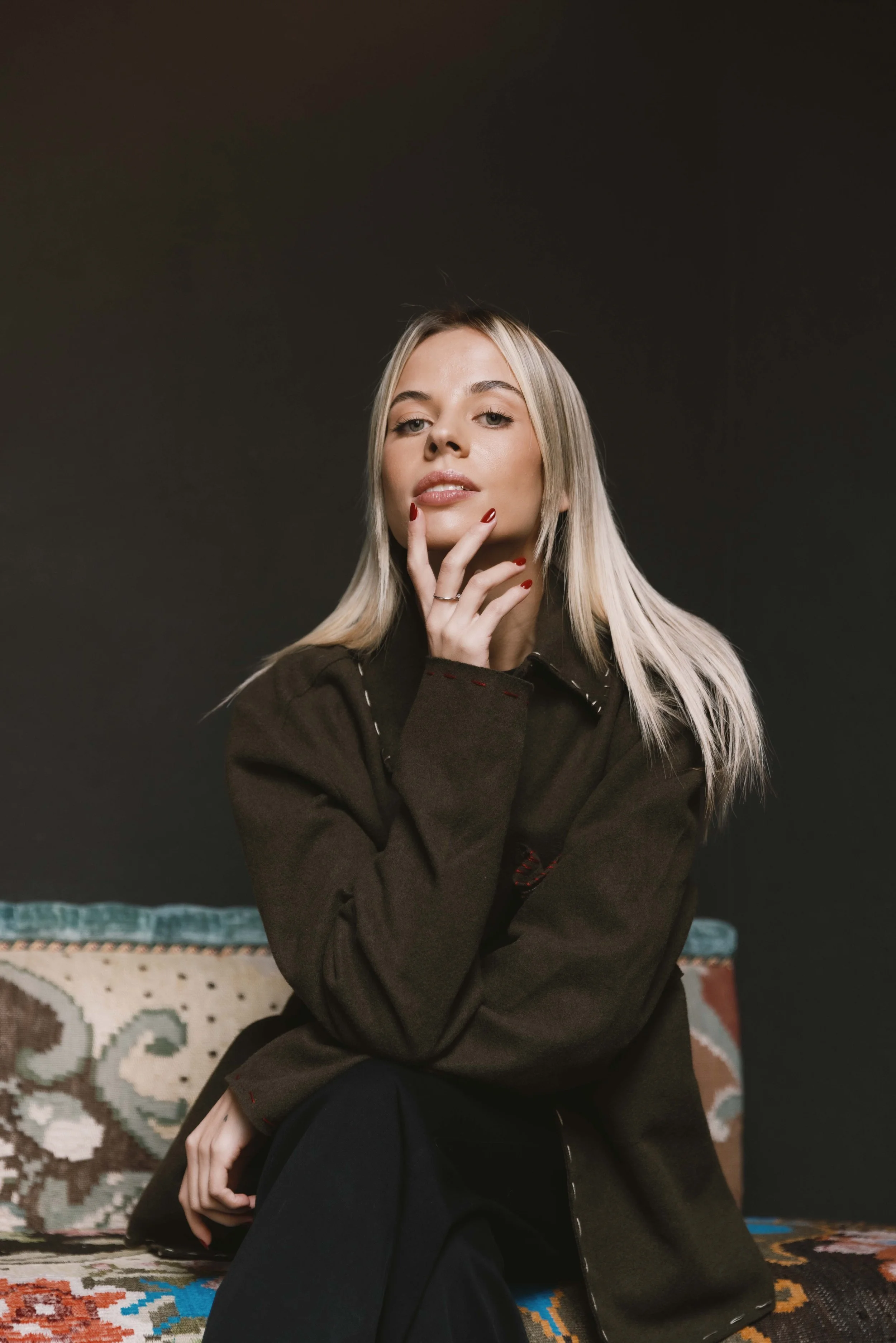 Una donna bionda seduta su un tappeto, indossa una giacca marrone e ha le unghie rosso sangue, con uno sfondo nero.