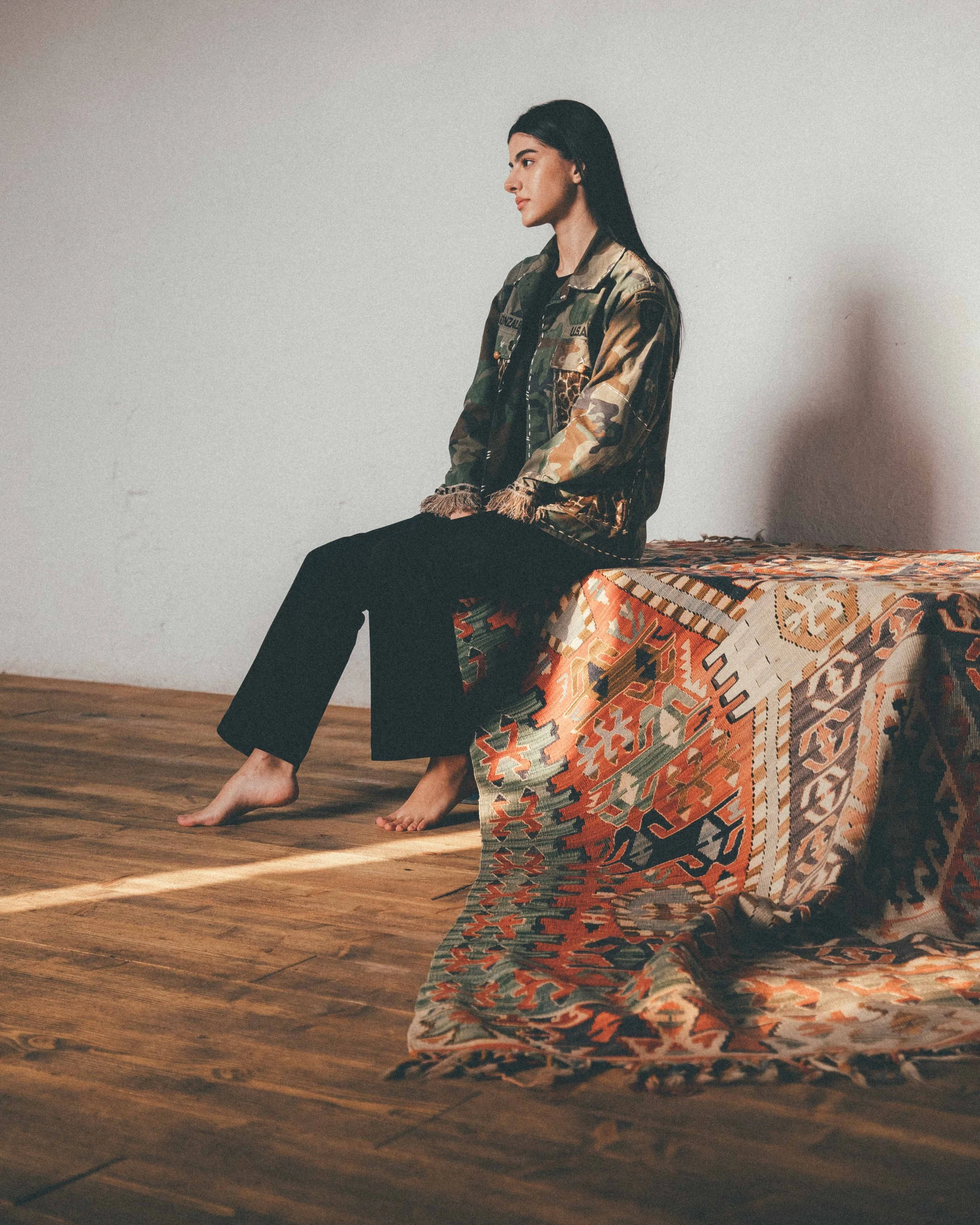 Ragazza seduta su una superficie coperta da un tappeto etnico, con parete bianca sullo sfondo, vestita con una giacca mimetica e pantaloni neri, con i piedi nudi