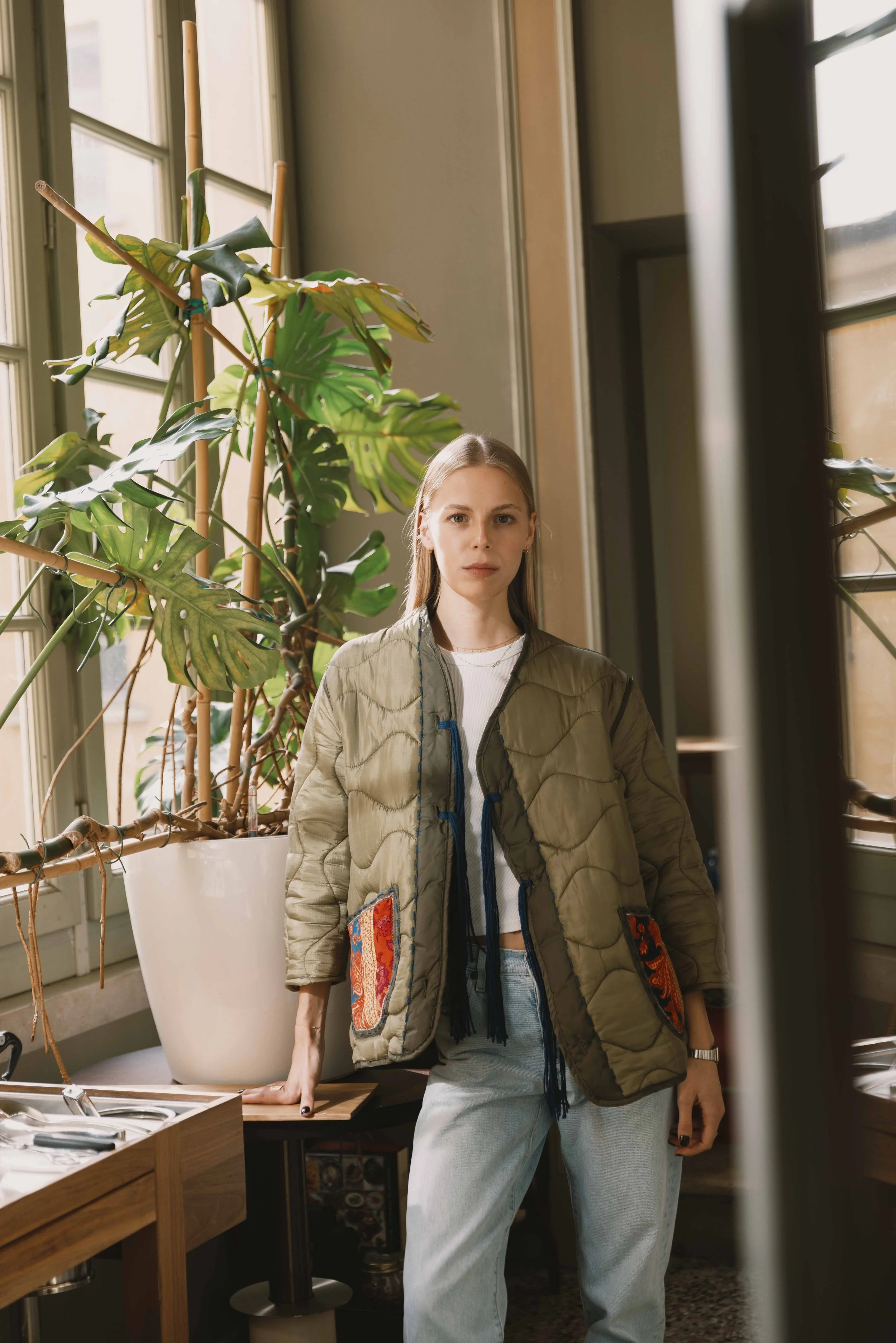 Una giovane donna con camicia bianca e pantaloni di jeans chiari si trova in una stanza con grandi finestre e una pianta tropicale. La donna porta una giacca verde con tasche decorative.