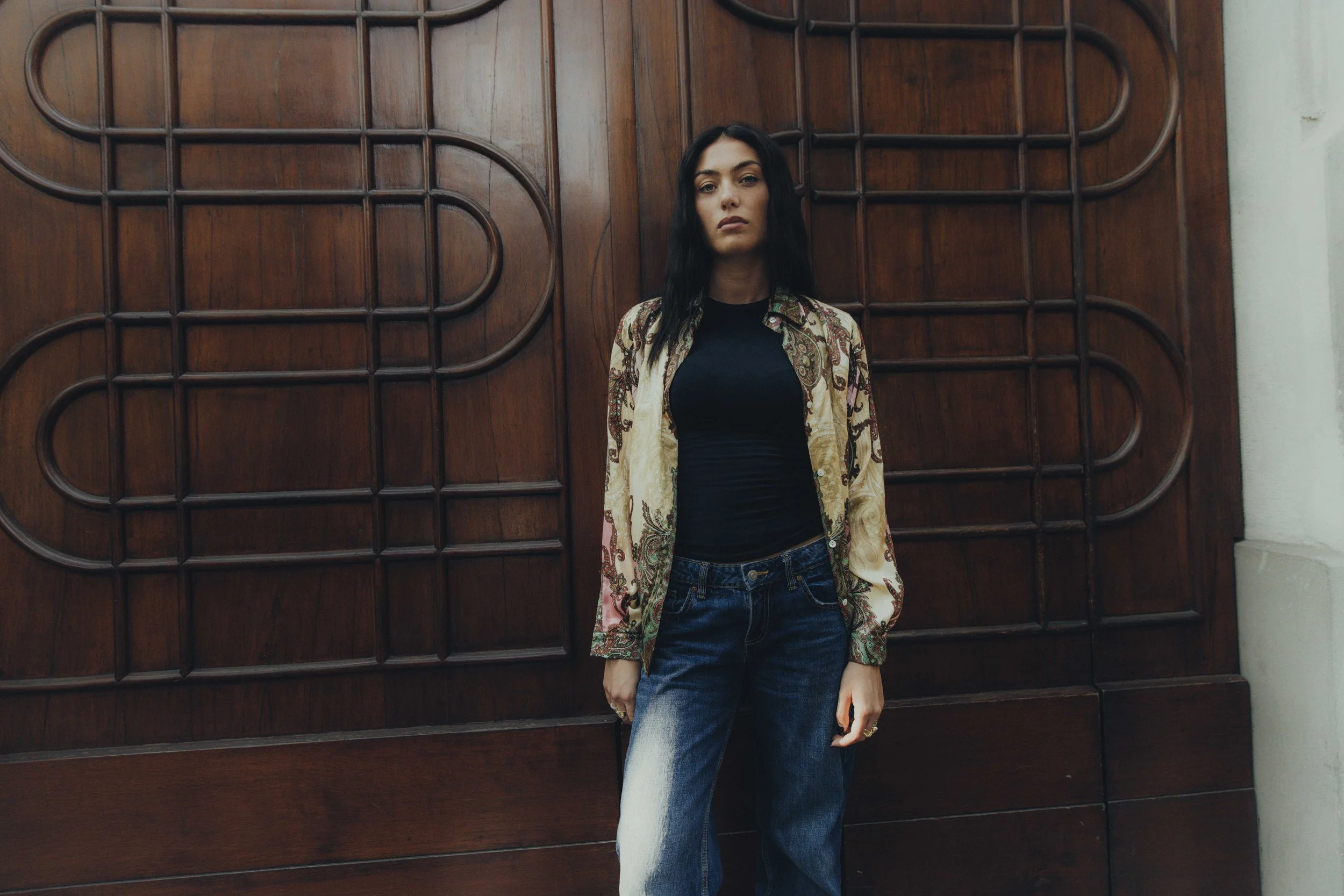 Ragazza con camicia colorata e pantaloni larghi davanti a una grande porta di legno