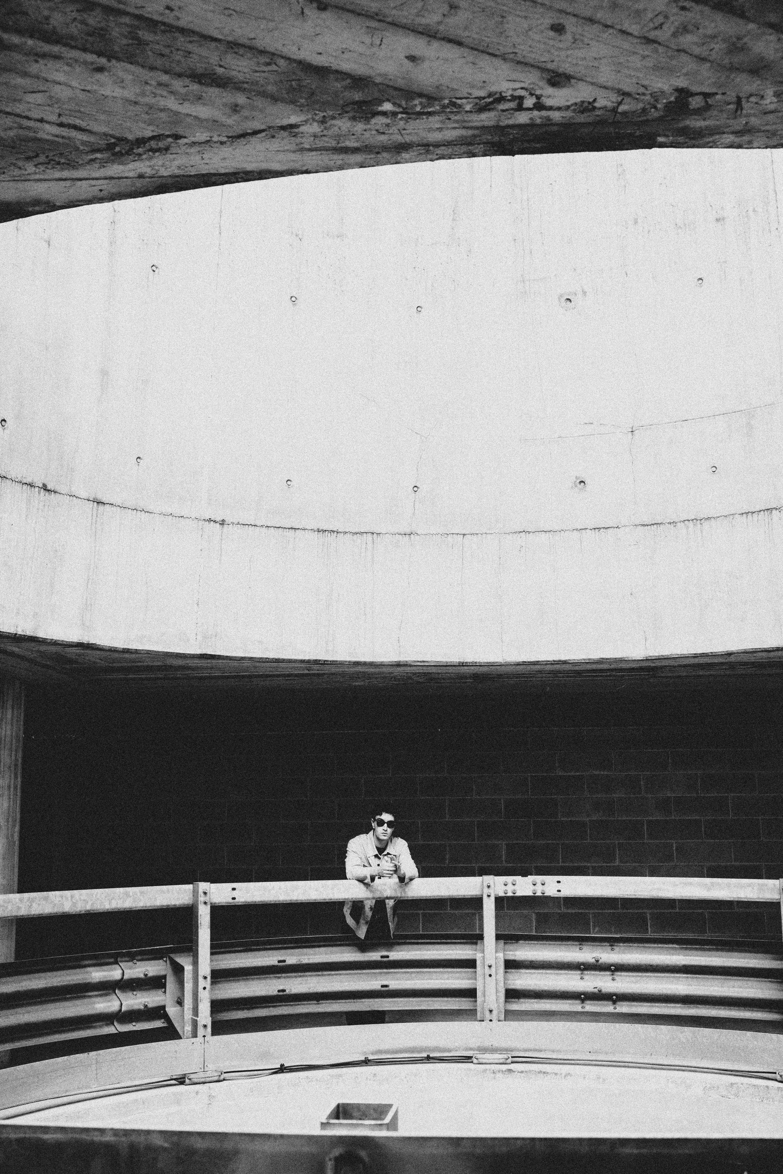 Un uomo con occhiali da sole si trova su una balconata in un edificio moderno con pareti scure e un soffitto in cemento, sullo sfondo una parete bianca curva
