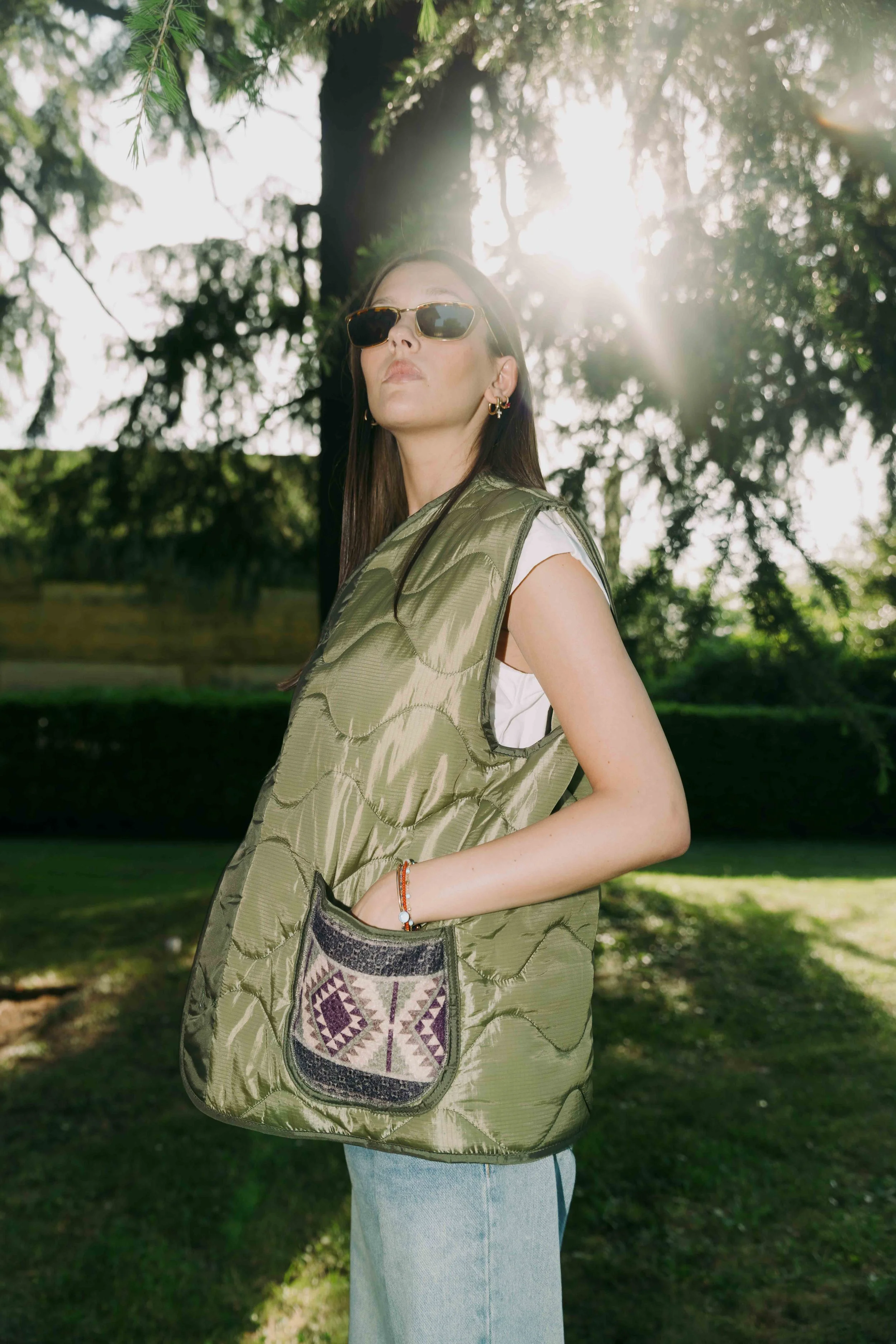 Donna con vestito estivo, occhiali da sole e giacca con tasca decorata, in un parco con alberi e sole al tramonto.