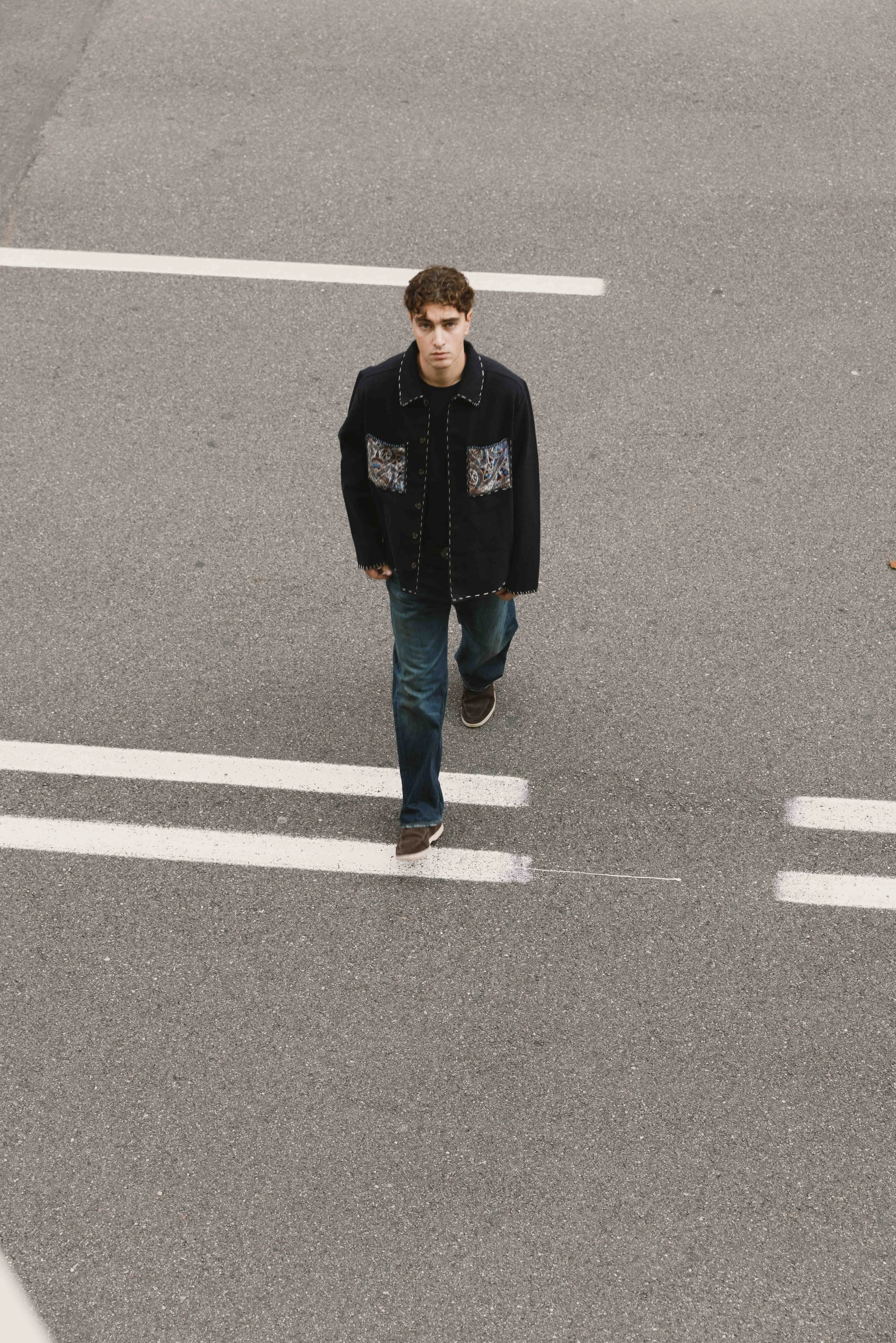 Un giovane uomo con giacca nera e jeans cammina in una strada vuota vista dall'alto.