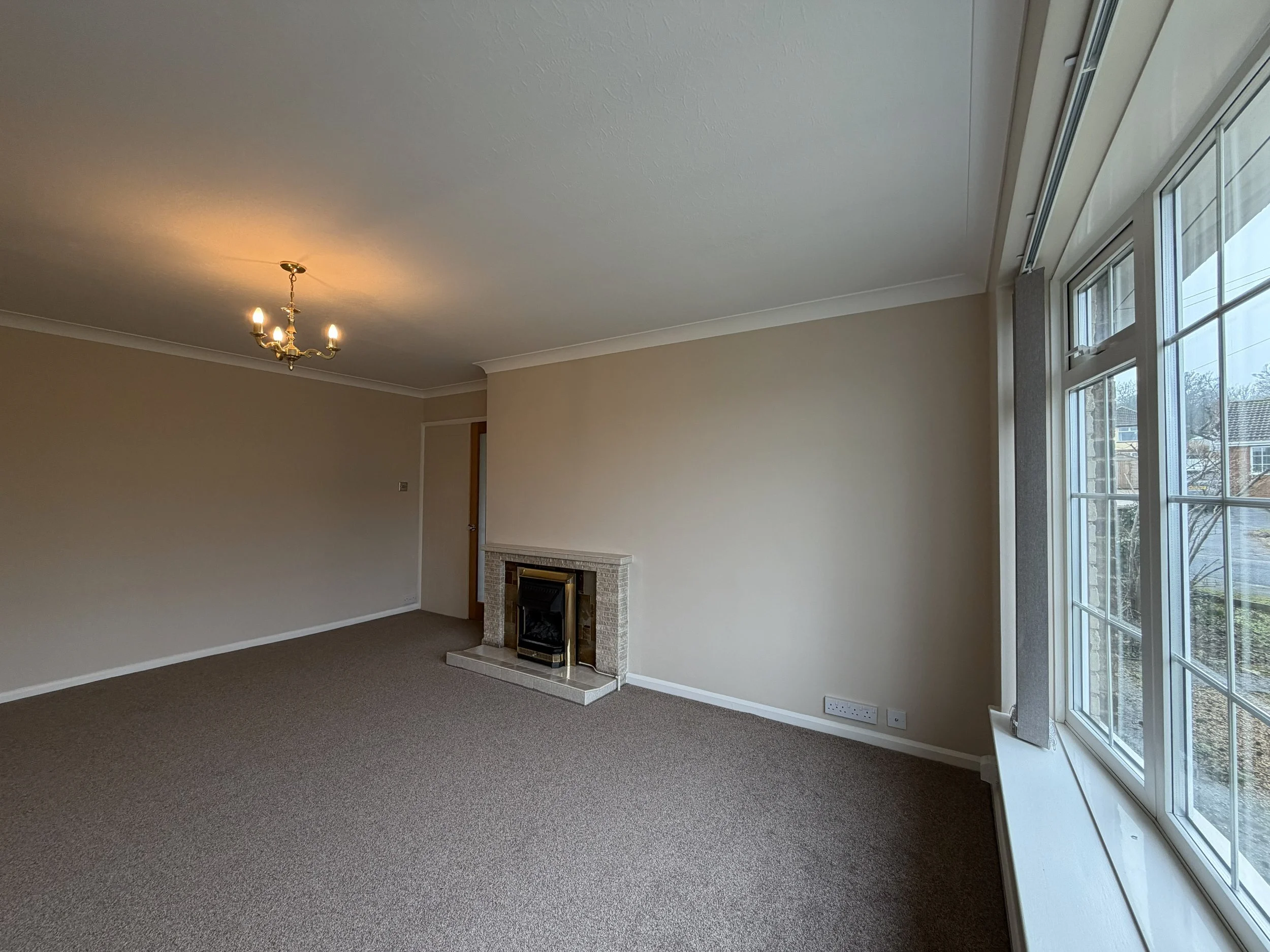 Empty living room with beige walls, a large window on the right, a small fireplace on the back wall, and a ceiling chandelier with four lights.