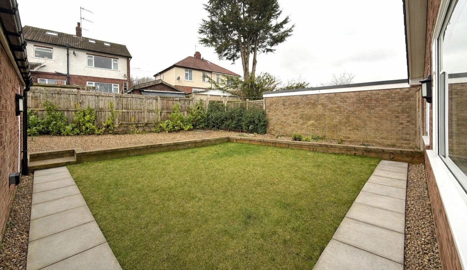 View of a backyard garden with a grassy lawn, bordered by stone pathways, a small raised flower bed with plants, and a brick wall fence with neighboring houses in the background.