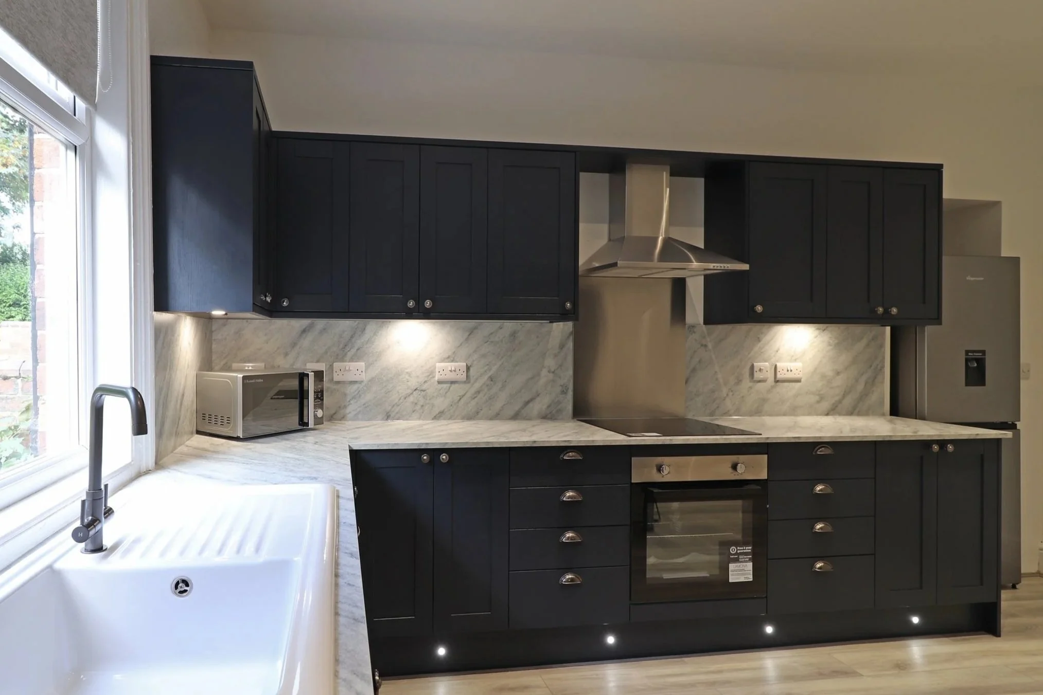 Modern kitchen with black cabinets, marble countertops, stainless steel appliances, and a kitchen sink by a large window.