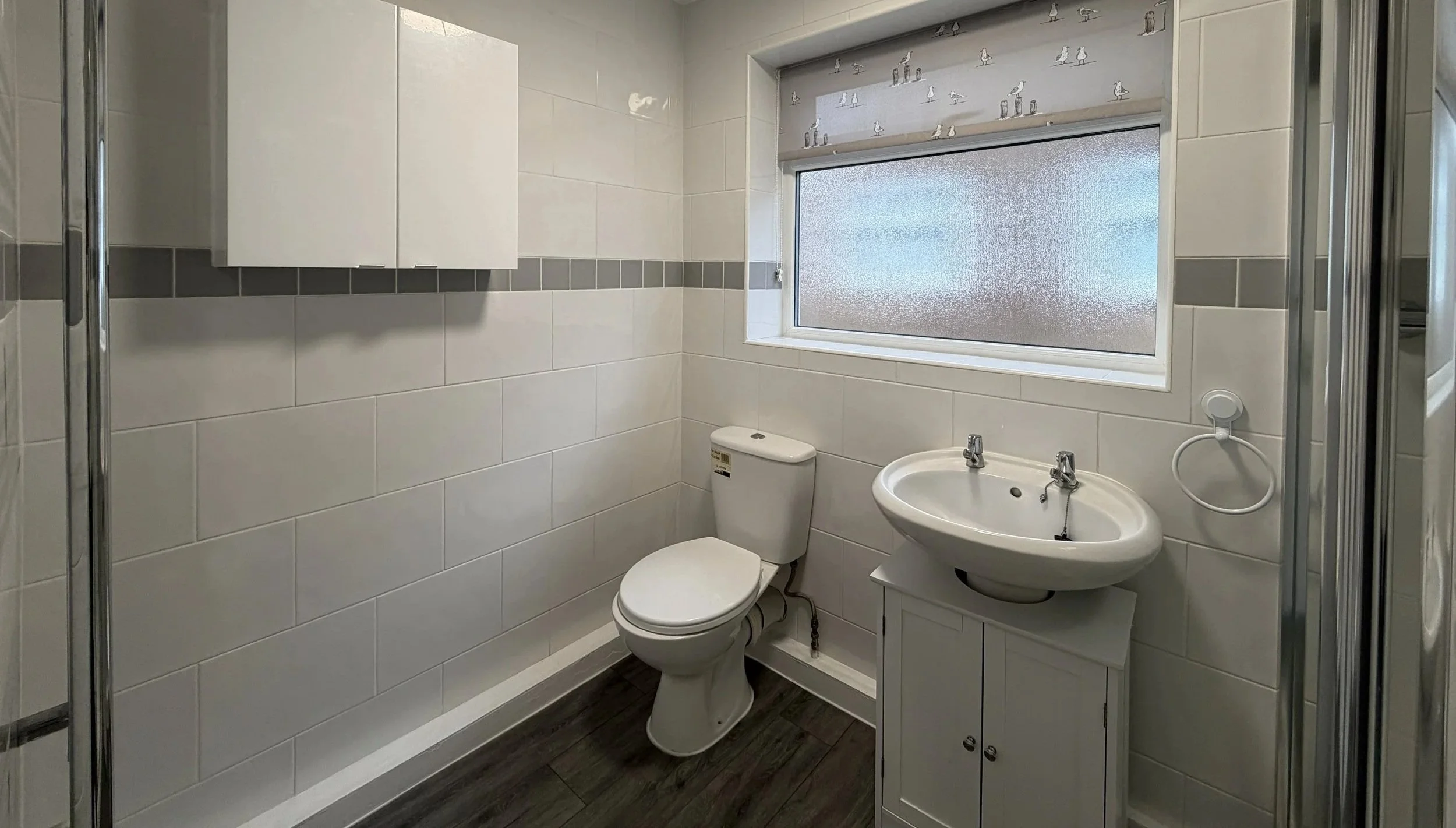 Bathroom with white tiles, a toilet, a small white cabinet with a sink and two faucets, a frosted window, and a patterned roller blind.