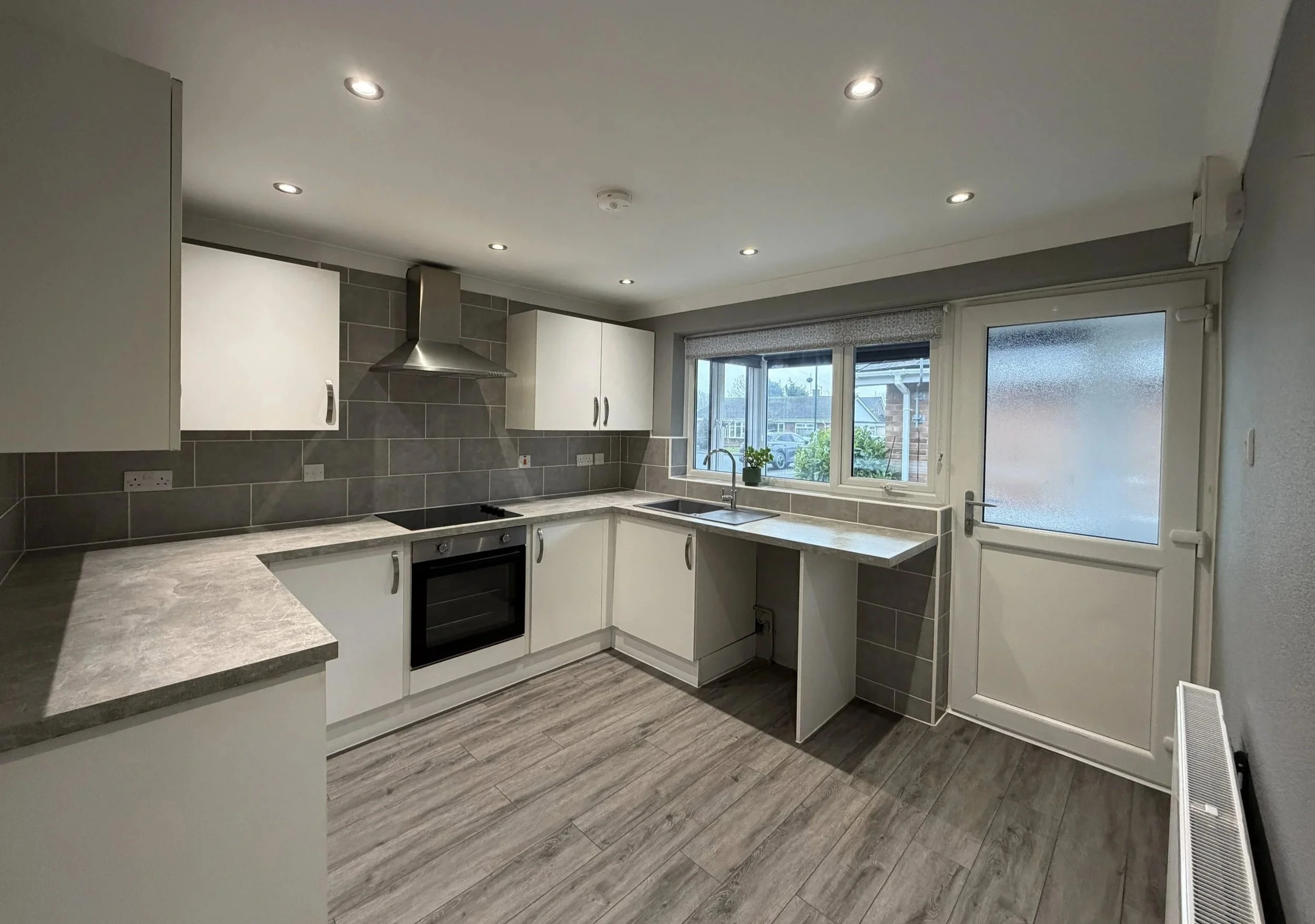 Modern kitchen with white cabinets, gray countertops, gray tiled backsplash, stainless steel range hood, built-in oven, sink under window, frosted glass door, wood-look flooring, ceiling recessed lighting, small potted plant on window sill.