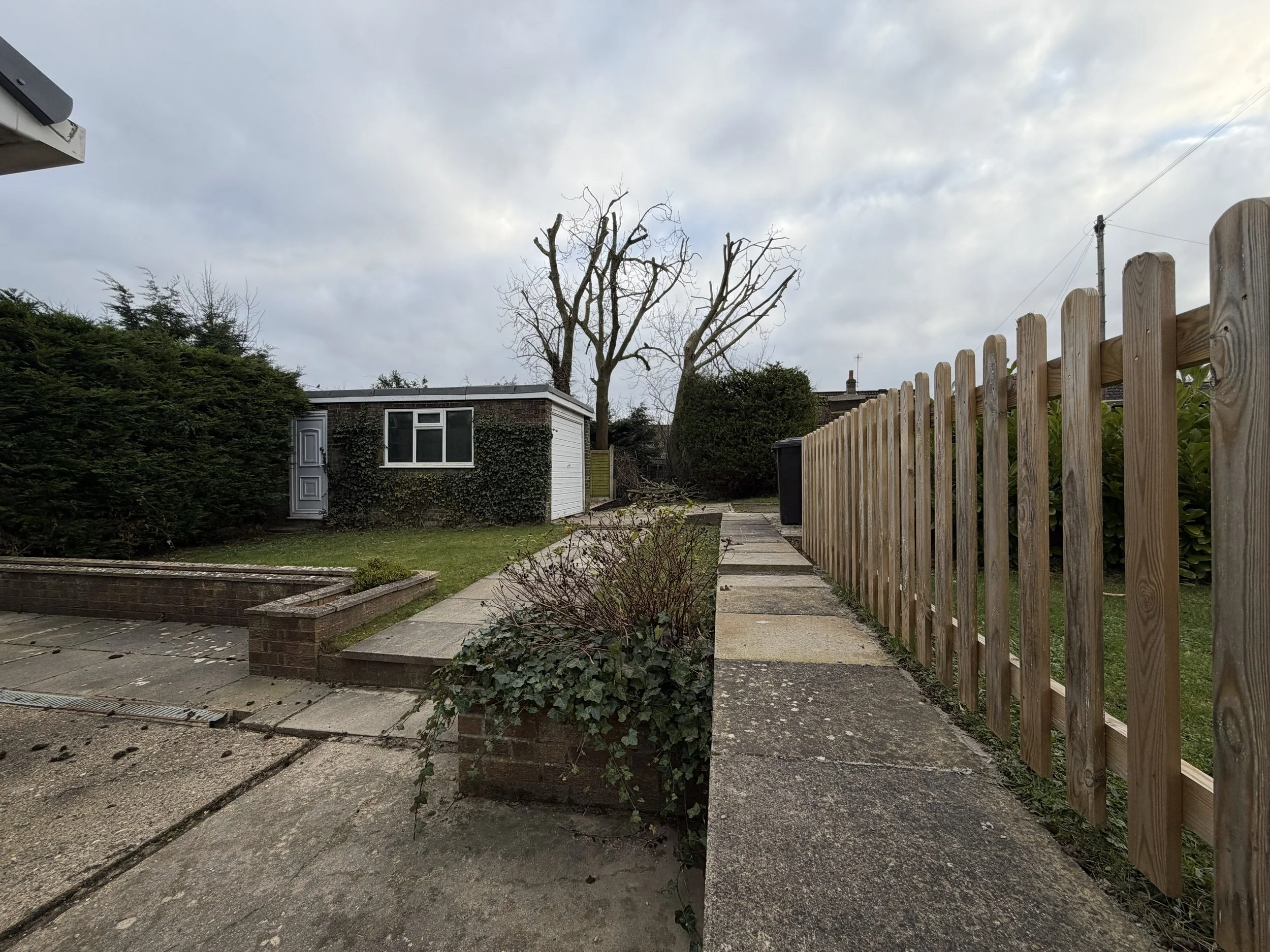 Backyard with concrete patio, flower bed, small green lawn, a white shed with windows and a door, bare trees, a tall wooden fence on the right, and cloudy sky.
