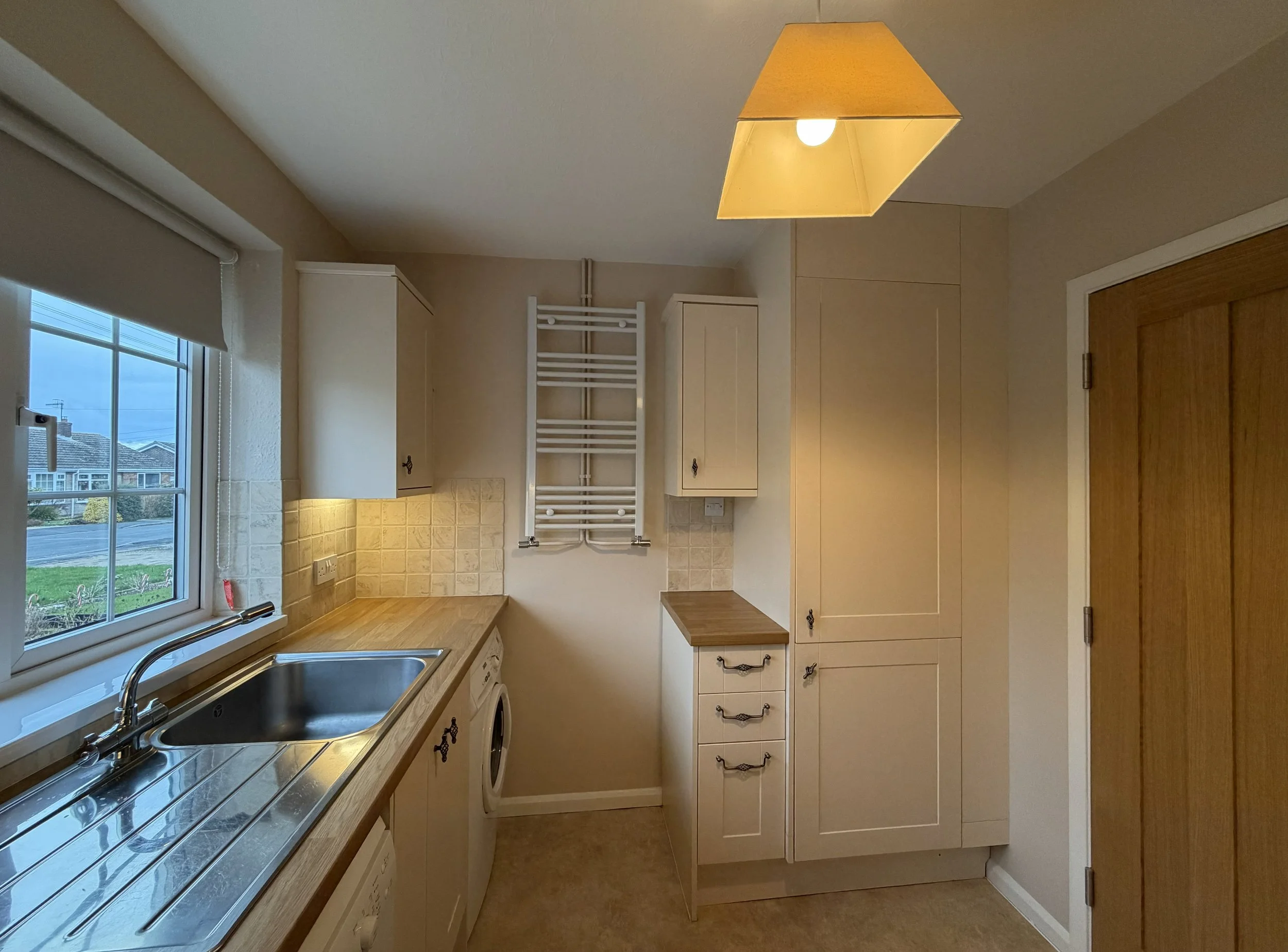 Kitchen with beige cabinets, wooden countertop, stainless steel sink, window, beige tiled backsplash, washing machine, white wall-mounted towel radiator, and closed wooden door, with a ceiling light fixture.