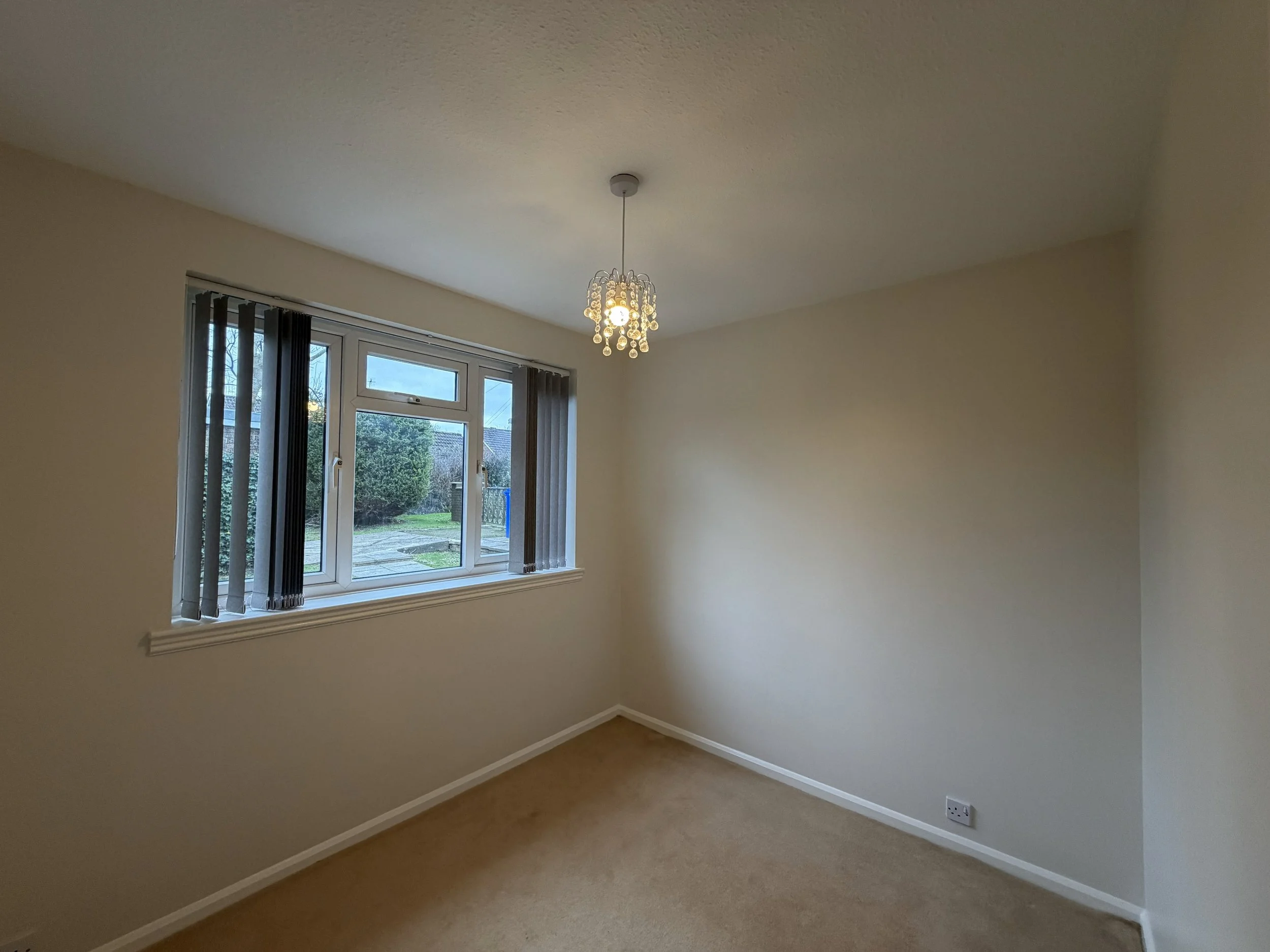 Empty room with beige carpet, cream-colored walls, a window with vertical blinds, and a ceiling light fixture with decorative hanging crystals.