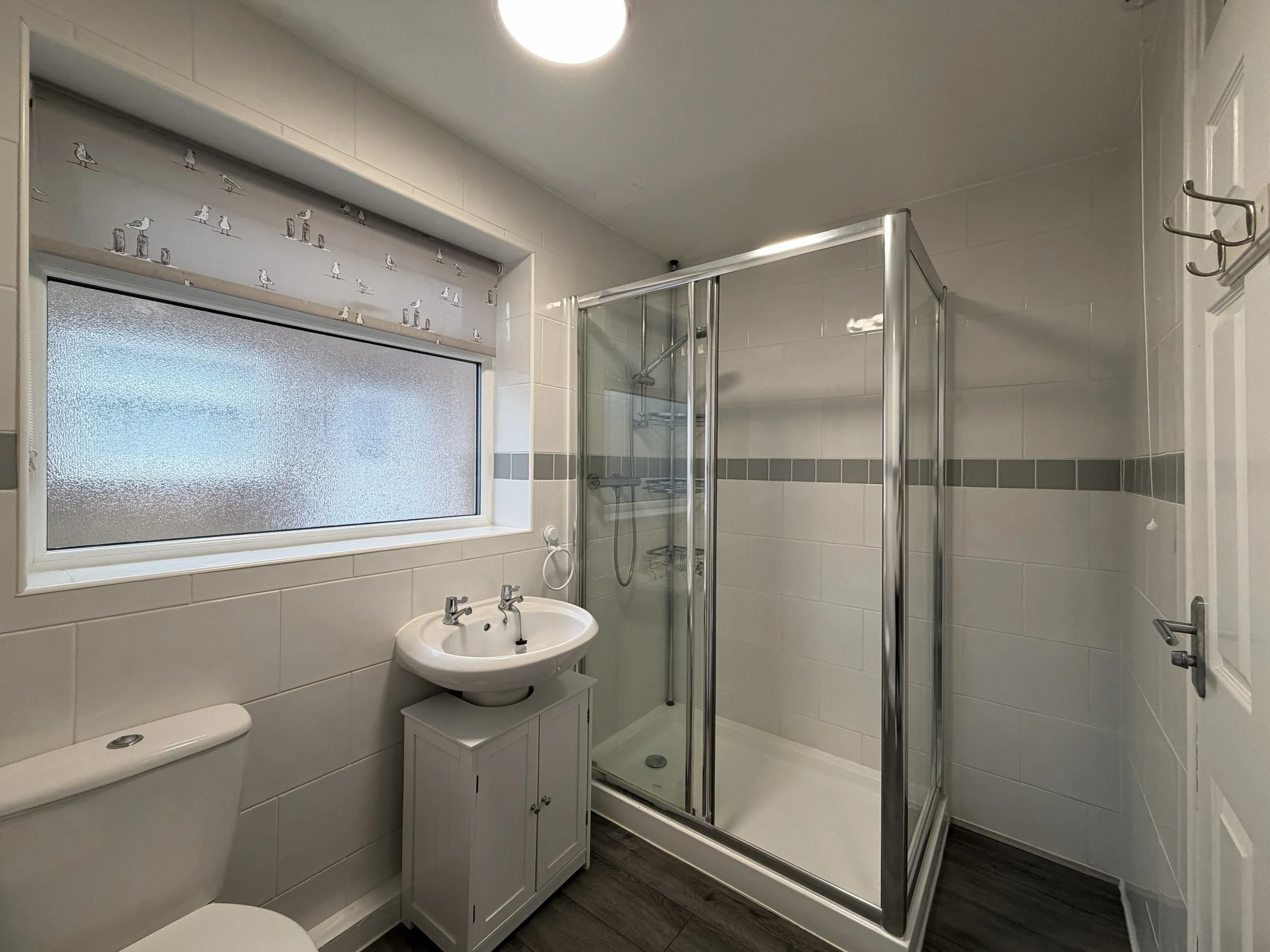 Bathroom with a frosted window, a white toilet, a small white sink cabinet, and a glass shower enclosure with tiled walls.