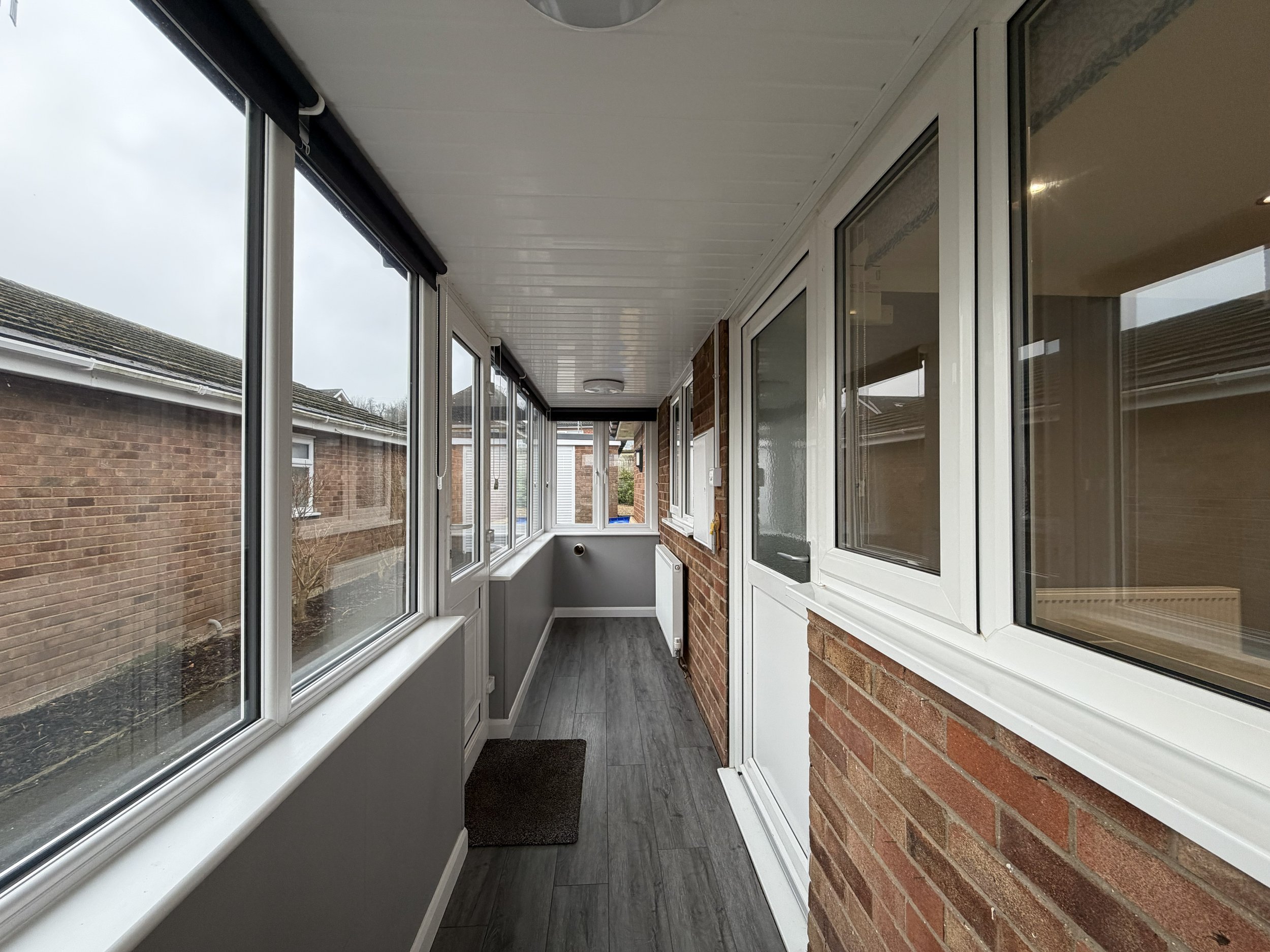 Enclosed porch with large windows, brick wall, dark wood flooring, and a small black doormat.