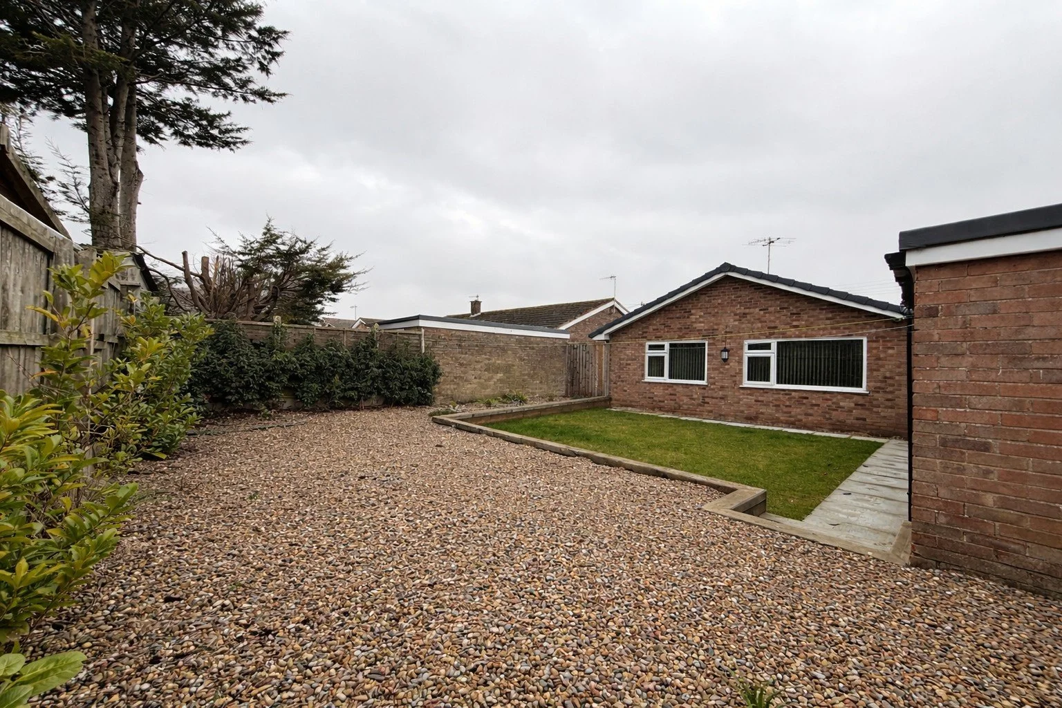 A backyard with a gravel area on the left, a small grass lawn on the right, and a brick house with three windows on the far side. There's a wooden fence on the left, bushes along the fence, and a cloudy sky overhead.