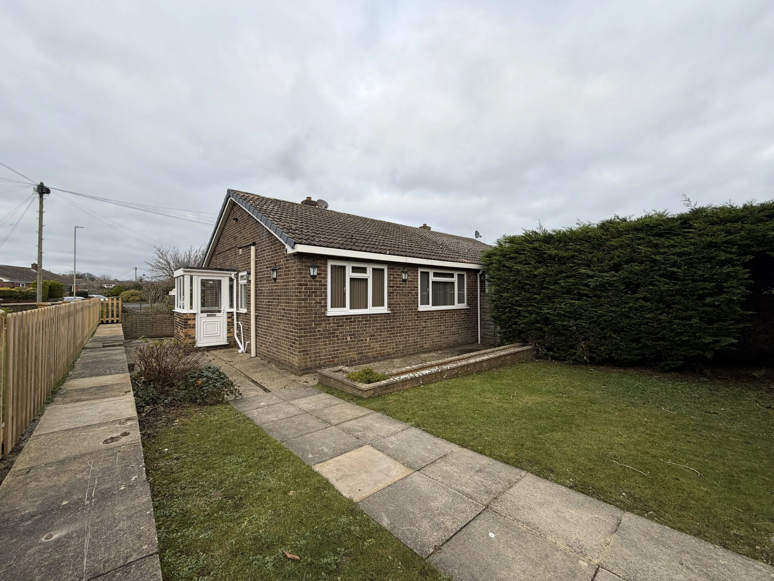 A small brick house with a gray roof, white window frames, and an enclosed porch. There is a lawn, a paved walkway, and a wooden fence on one side with a hedge on the other.