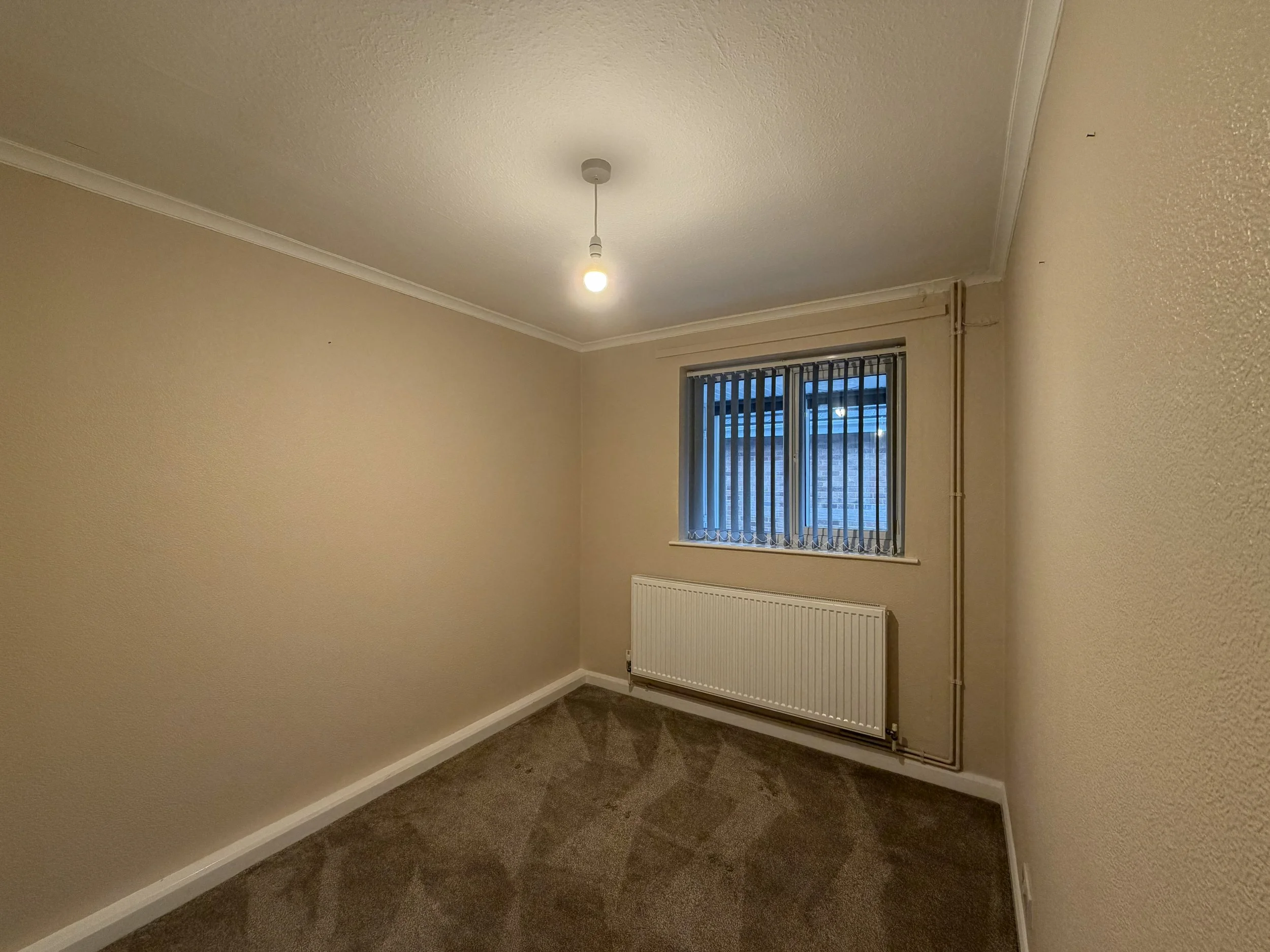 Empty small room with beige walls, brown carpet, a window with vertical blinds, a radiator below the window, and a ceiling light bulb.