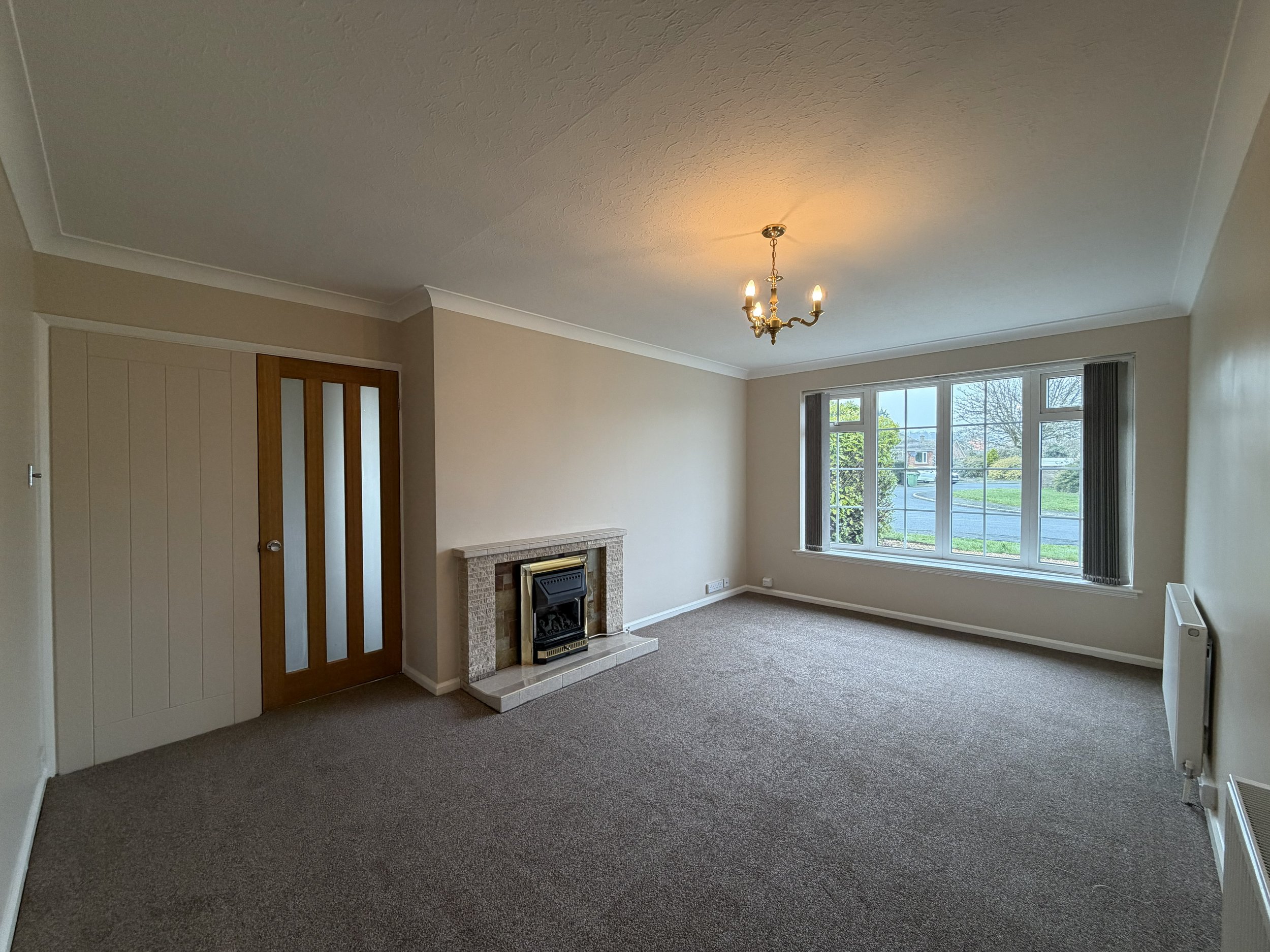 Empty living room with beige walls, large window with vertical blinds, carpeted floor, small fireplace, and ceiling light fixture.