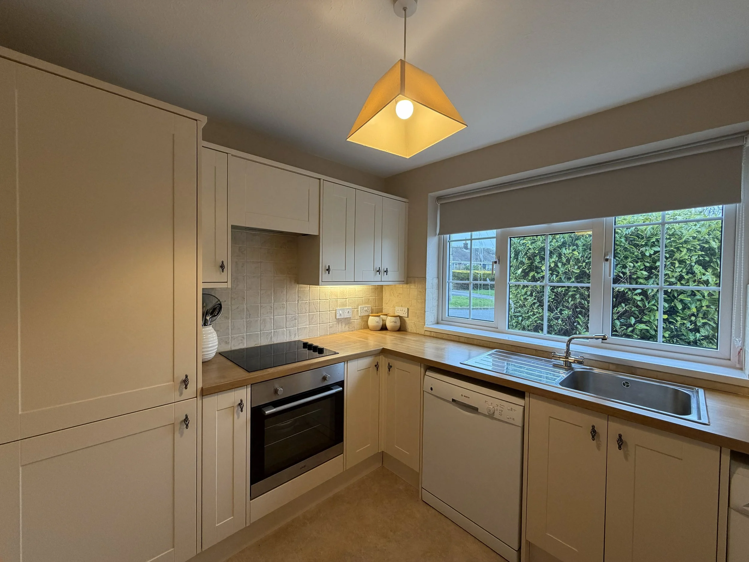 Kitchen with cream-colored cabinets, wooden countertop, black electric stove, oven, dishwasher, and a stainless steel sink beneath a large window showing green foliage outside, illuminated by a hanging geometric pendant light.