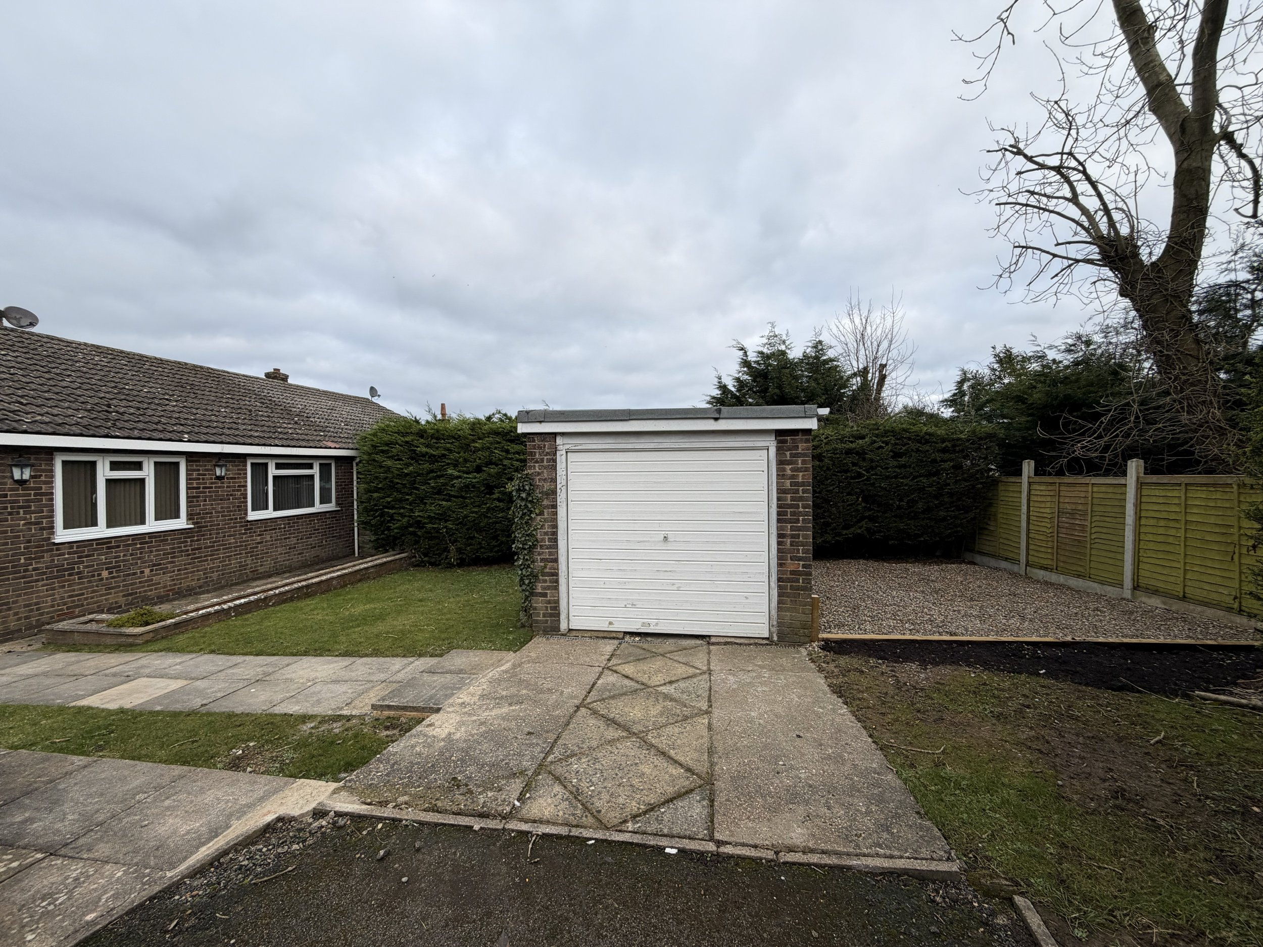 Backyard with concrete pathway leading to a small brick garage with white door, to the right is a gravel area, and to the left are green lawns and a brown brick house with white-framed windows, trees and a cloudy sky.