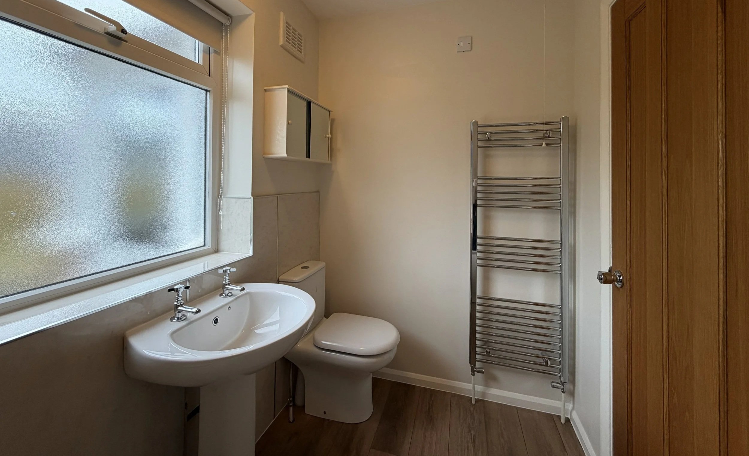 A small bathroom with a frosted window, a white sink with chrome taps, a toilet, a small white cabinet, a chrome towel radiator, and a wooden door.