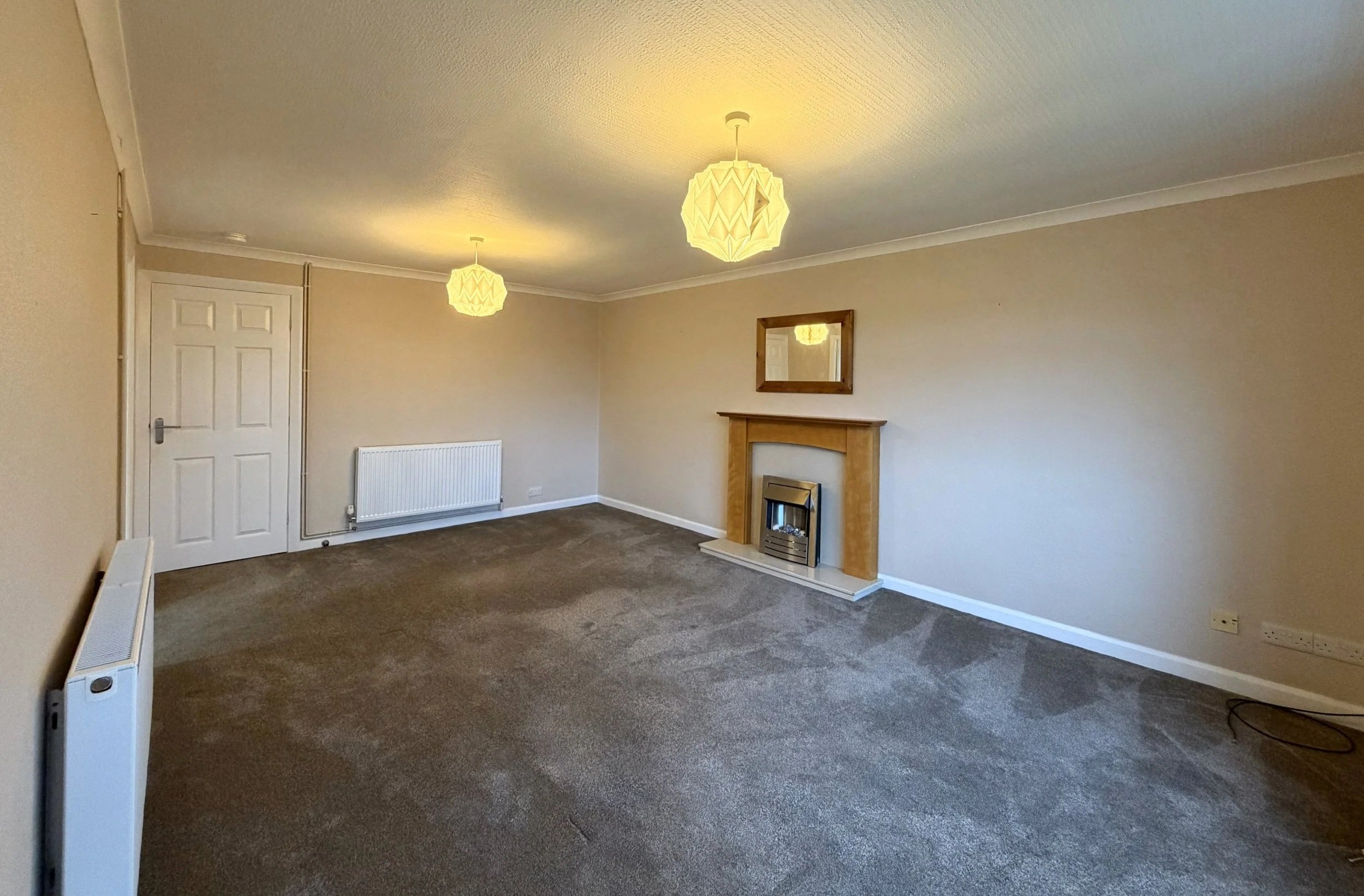 Empty living room with beige walls, brown carpet, a fireplace with a wooden mantel, a mirror above the fireplace, two ceiling lights, a white radiator on the left wall, and multiple electrical outlets.
