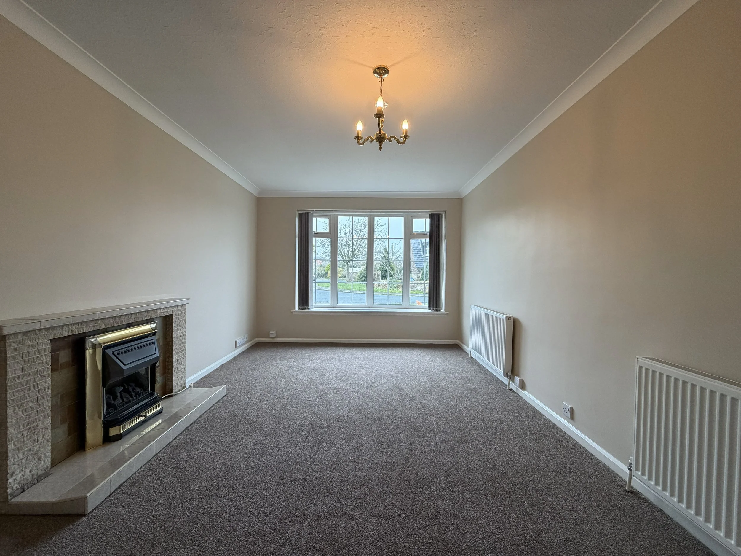 Empty living room with a large window, a fireplace, beige walls, a chandelier, and brown carpet.