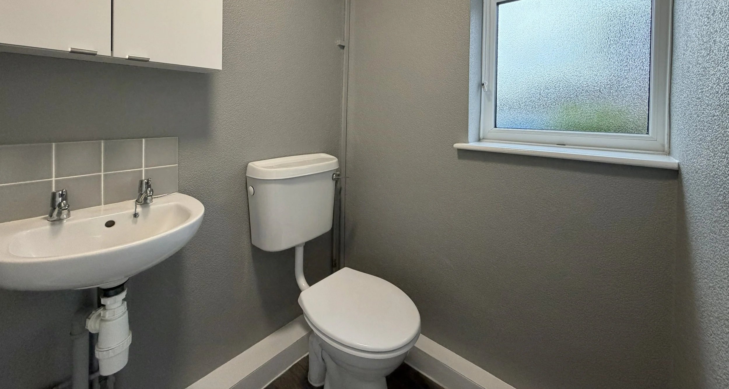 Small bathroom with a white toilet, wall-mounted sink, window with frosted glass, gray textured walls, and dark wood flooring.