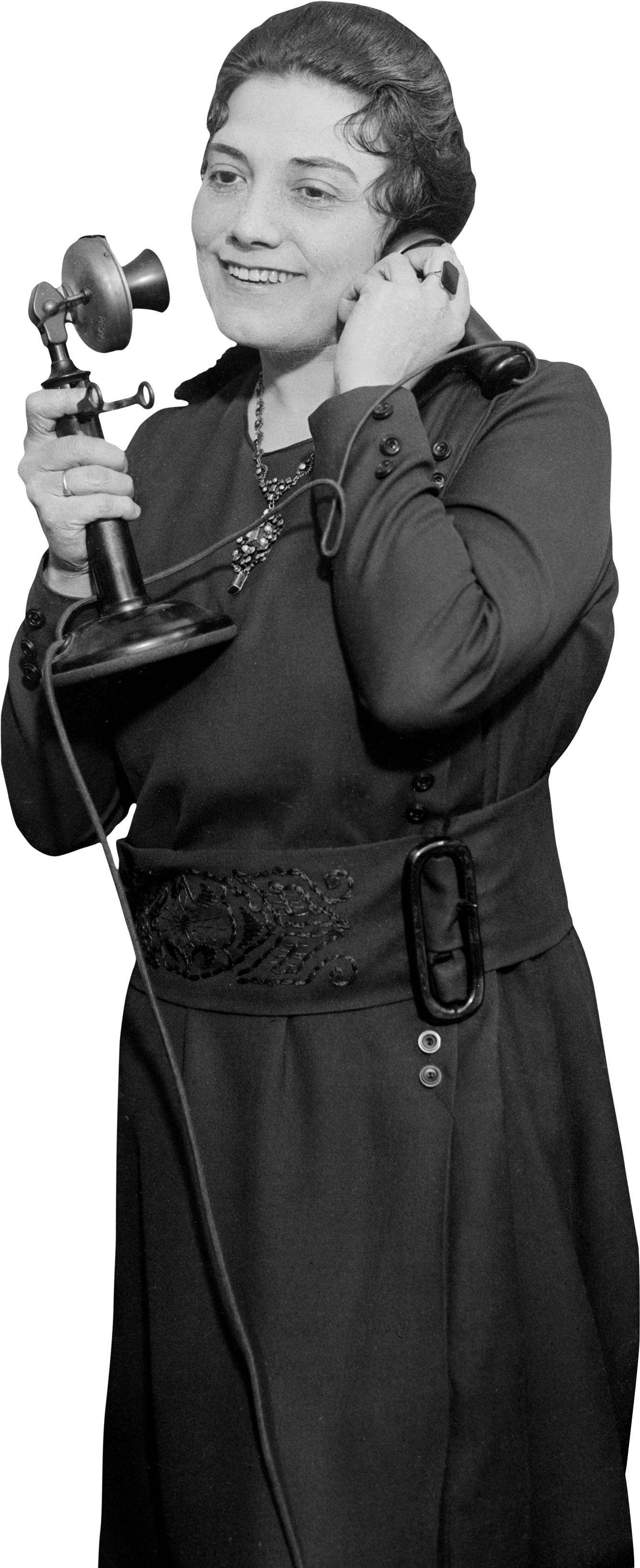 Black and white photo of a woman smiling while talking on a corded telephone, holding the receiver to her ear with her right hand and the phone base with her left hand. She is wearing a dark dress or blazer, a necklace, and a large belt with a decorative buckle.