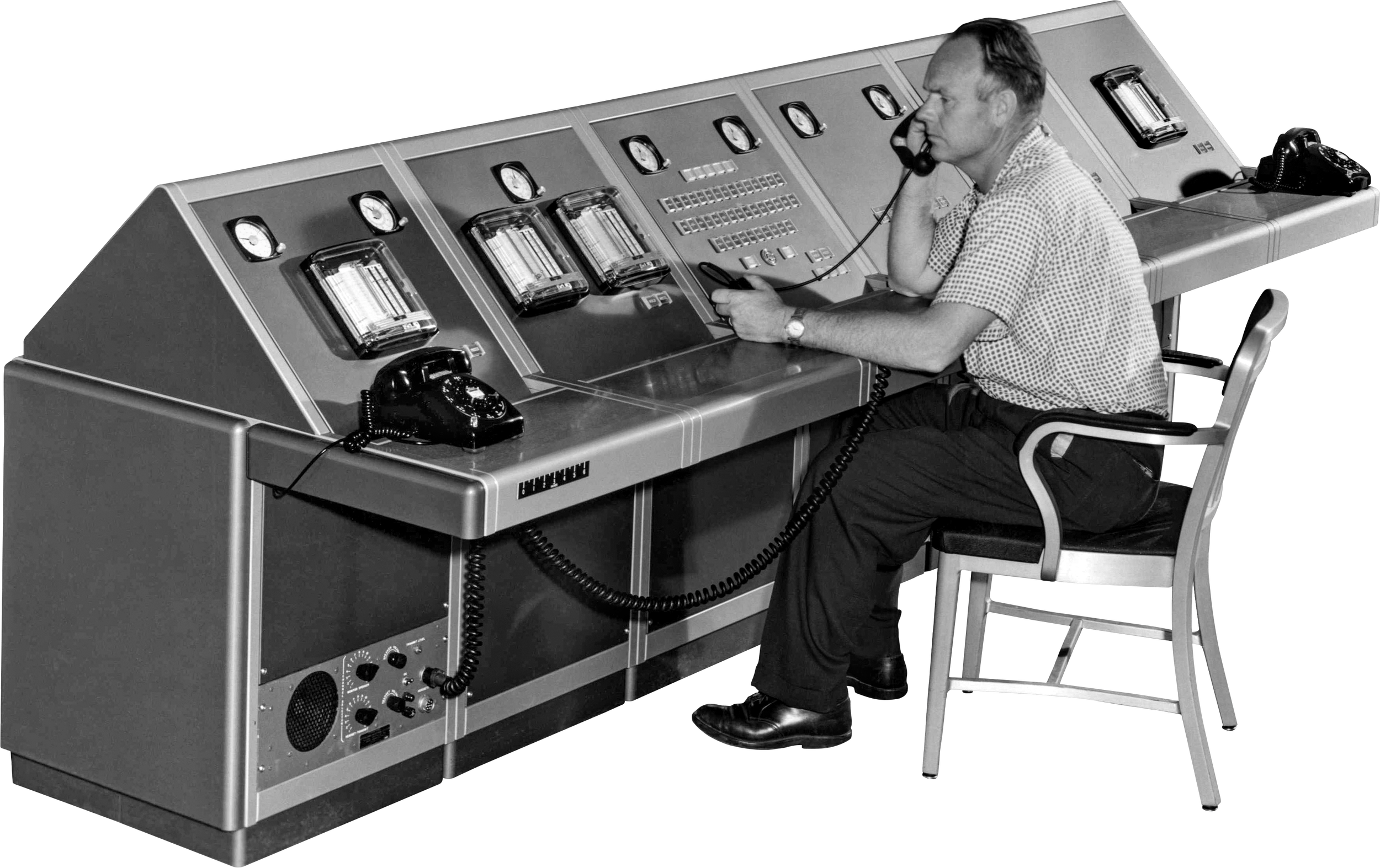 A man in a checkered shirt talking on a telephone at a large vintage control panel with dials and switches, holding a remote control device.