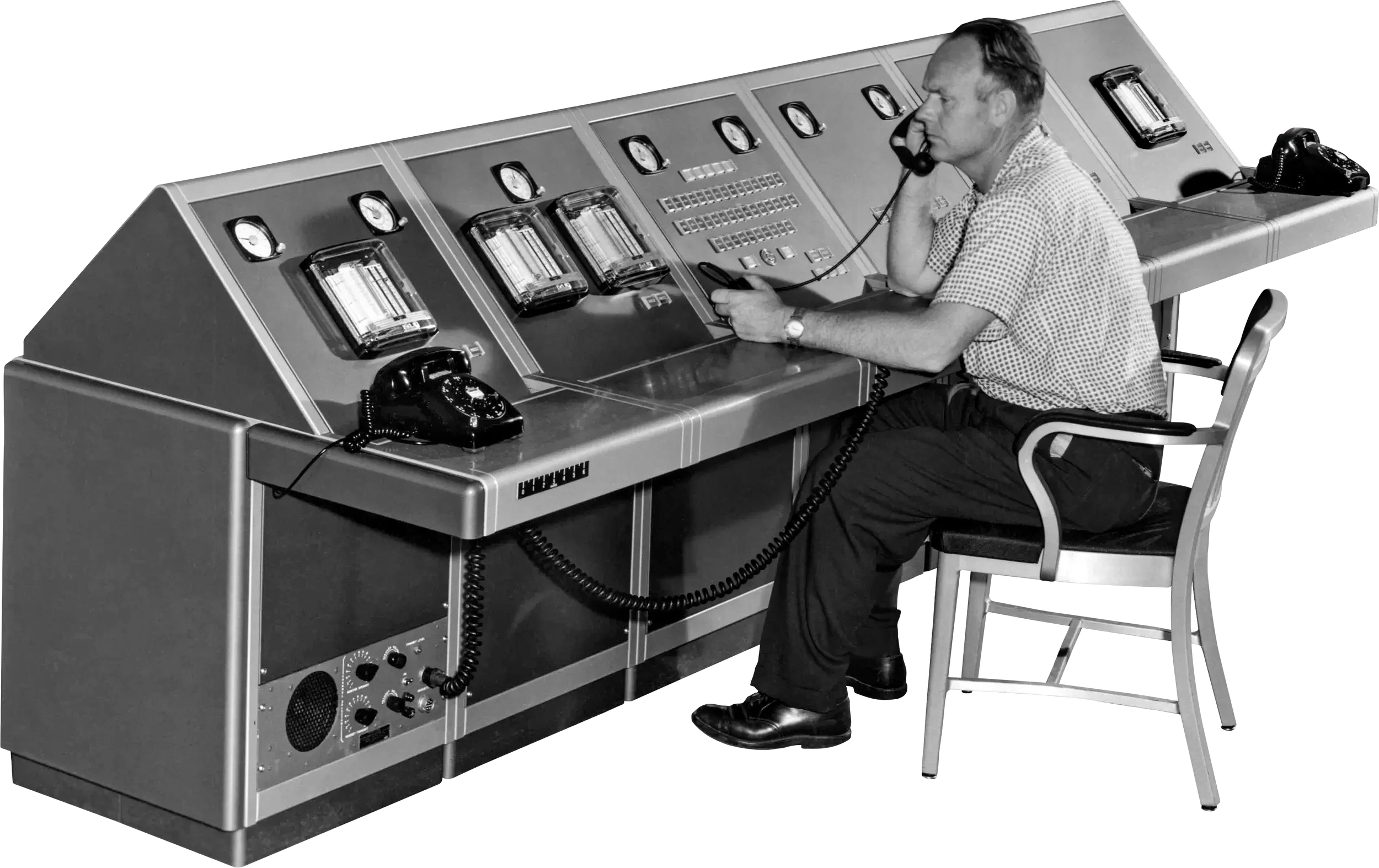 A man in a checkered shirt talking on a telephone at a large vintage control panel with dials and switches, holding a remote control device.