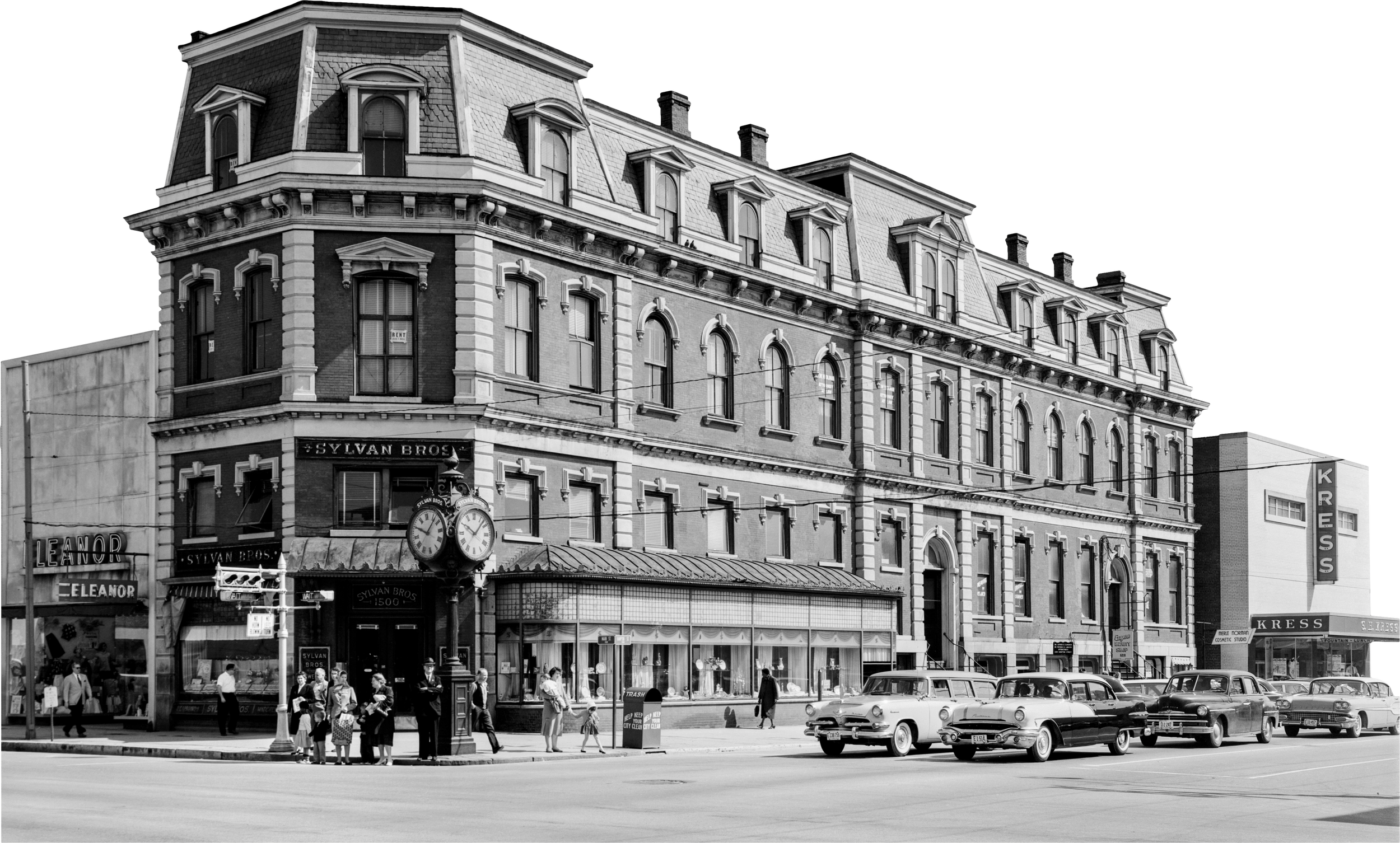 Black and white photo of a historic urban street scene with a large ornate brick building, classic cars parked in front, and people crossing the street.