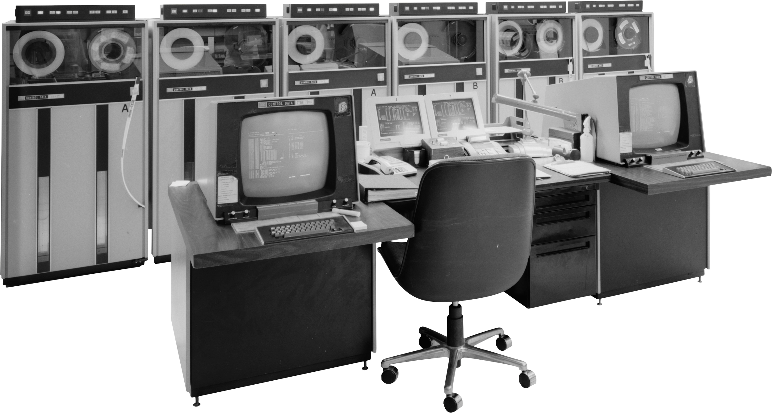 Black and white photo of a vintage control room with multiple old computer monitors, terminals, and telephones, along with a black office chair in front of the desks.