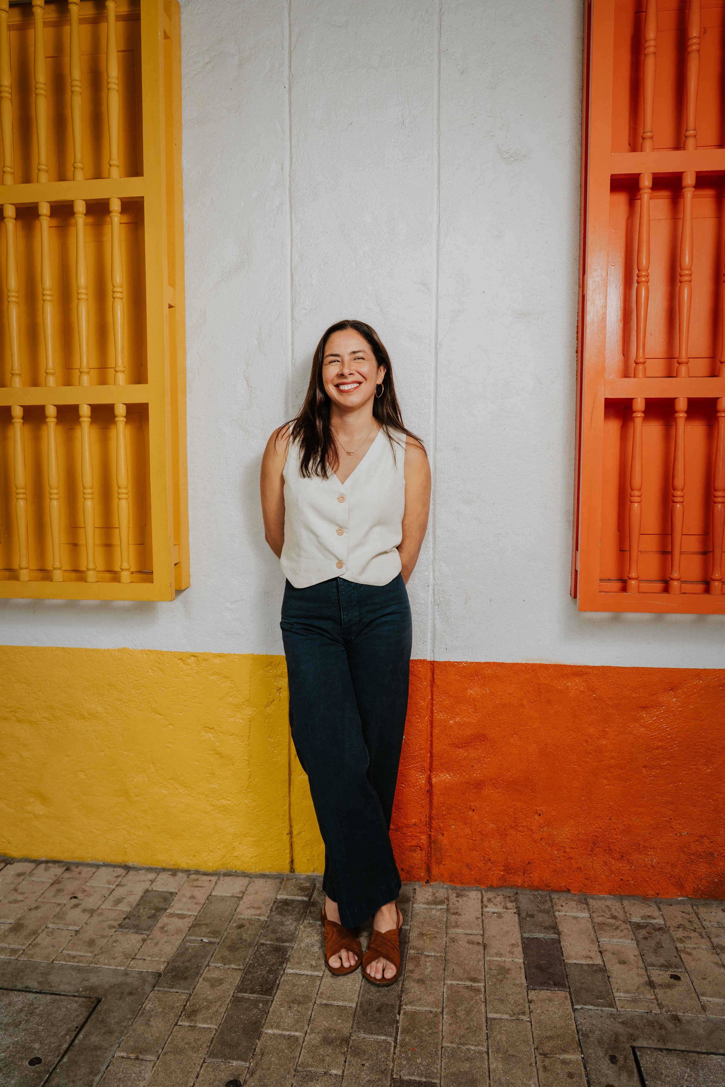 A woman with long dark hair smiling, standing with her back against a white wall with yellow and orange wooden window shutters on either side, wearing a sleeveless beige vest, dark jeans, and brown sandals.