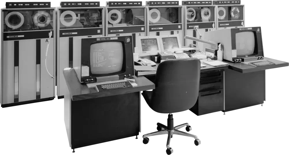 Black and white photo of a vintage control room with multiple old computer monitors, terminals, and telephones, along with a black office chair in front of the desks.